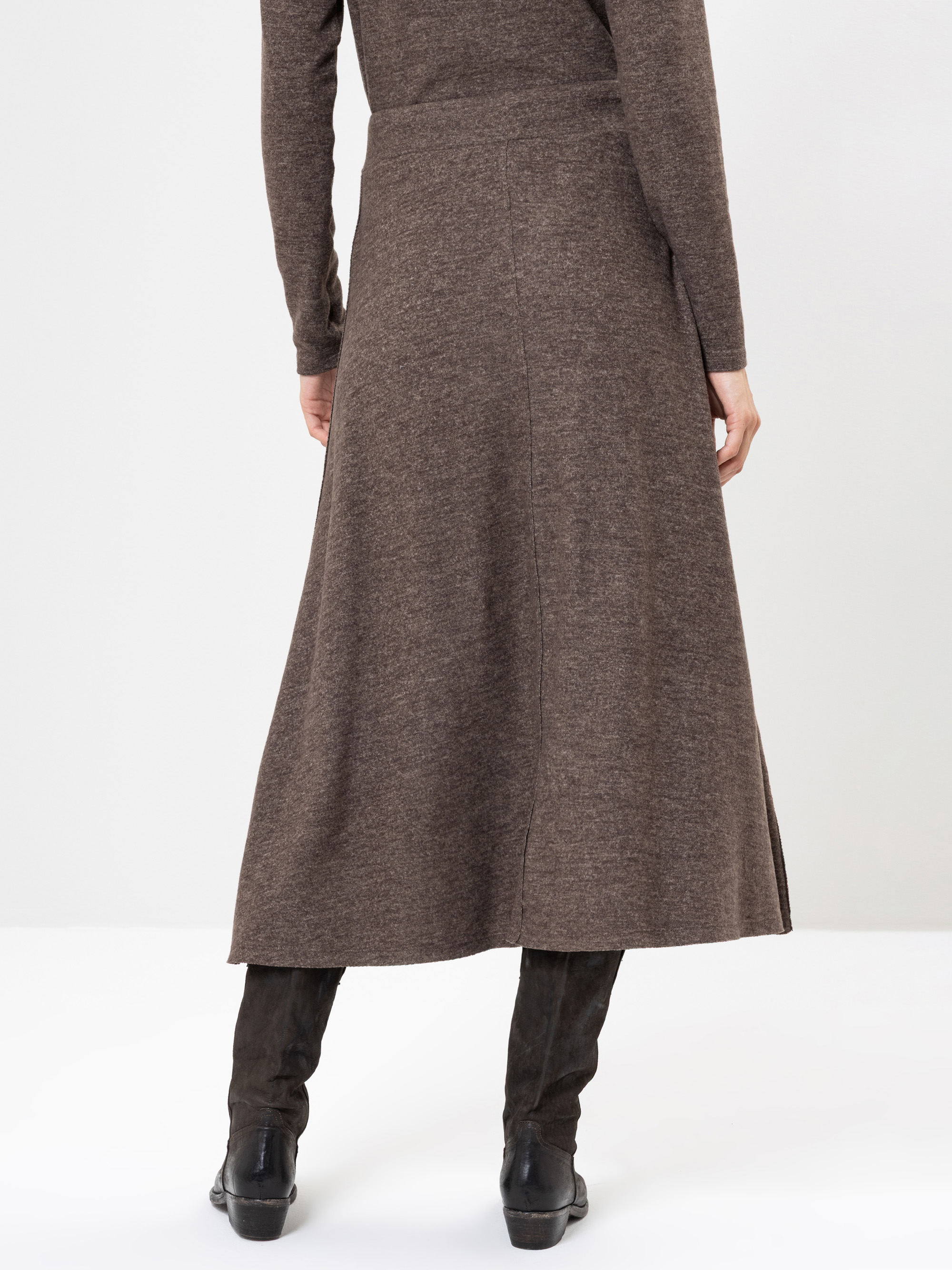 Eine Person steht mit dem Gesicht zur Wand, trägt ein braunes, langärmeliges Midikleid und schwarze, kniehohe Stiefel vor einem schlichten, hellen Hintergrund.