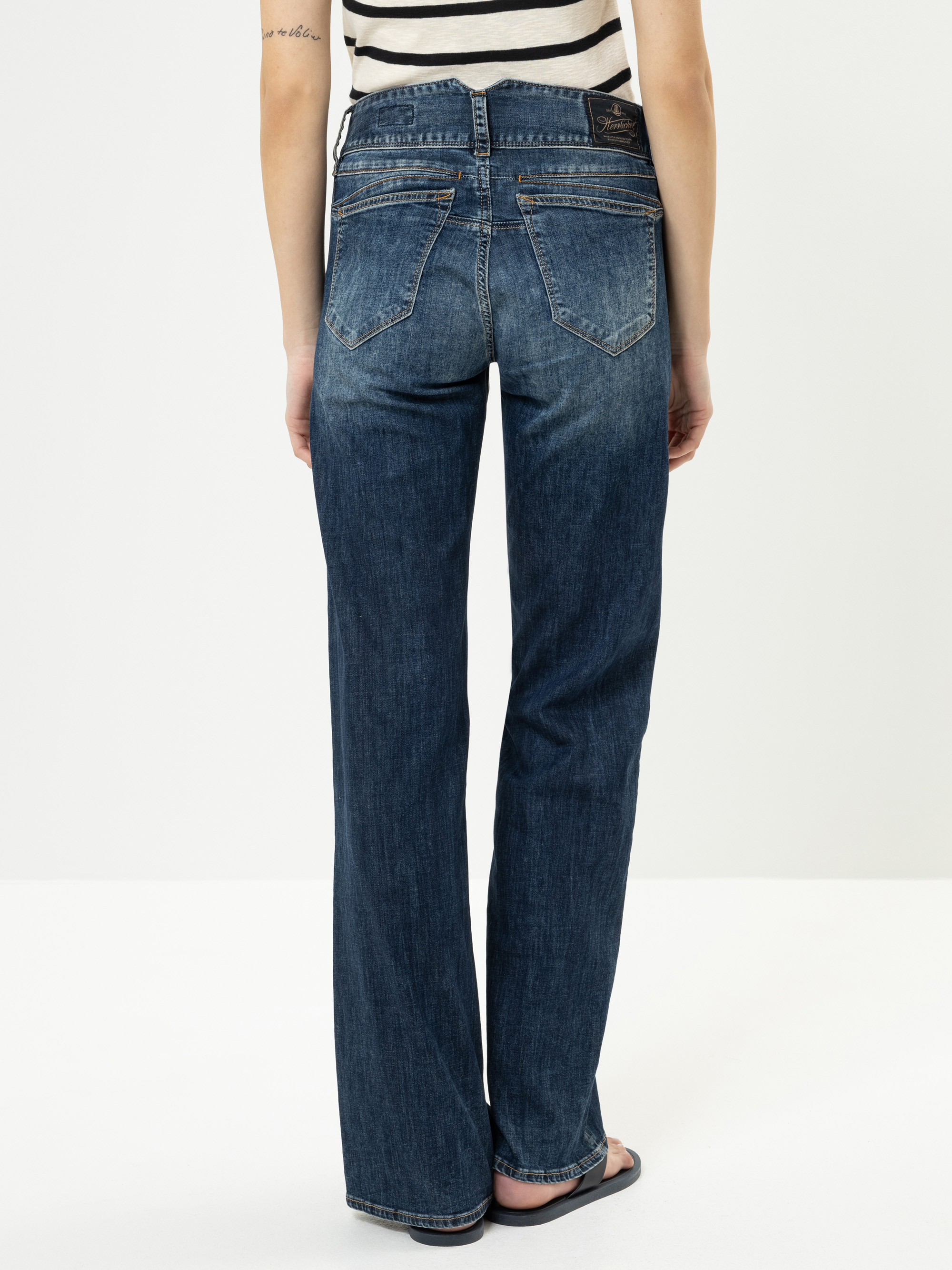 Eine Person, die blaue Jeans mit geradem Bein, ein schwarz-weiß gestreiftes ärmelloses Oberteil und schwarze Sandalen trägt, steht mit dem Gesicht zur Kamera vor einem schlichten weißen Hintergrund.