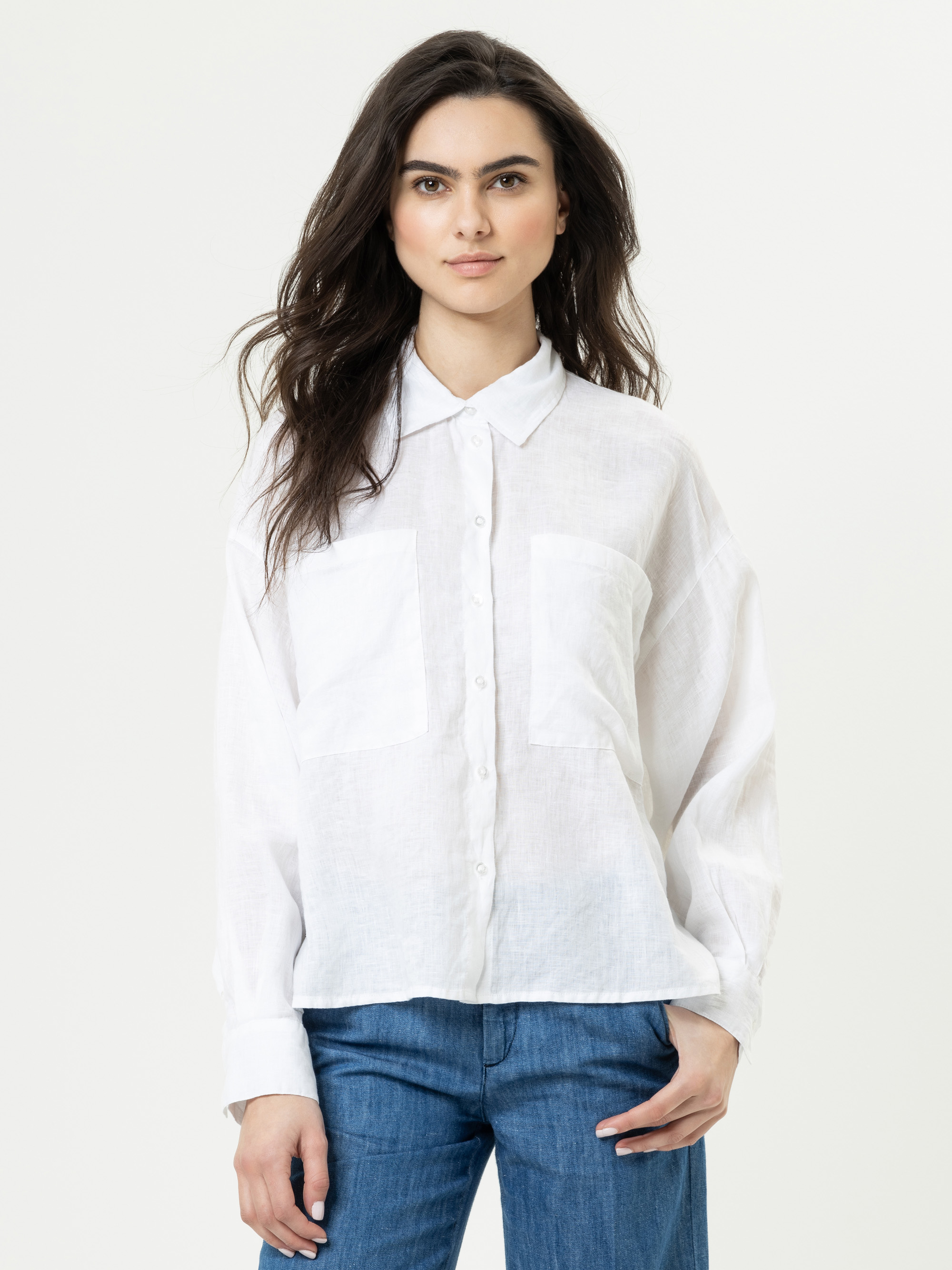 Eine Frau mit langen dunklen Haaren, die ein weißes Button-up-Hemd mit Vordertaschen und eine blaue Jeans trägt, steht vor einem schlichten hellen Hintergrund und blickt mit neutralem Blick in die Kamera.
