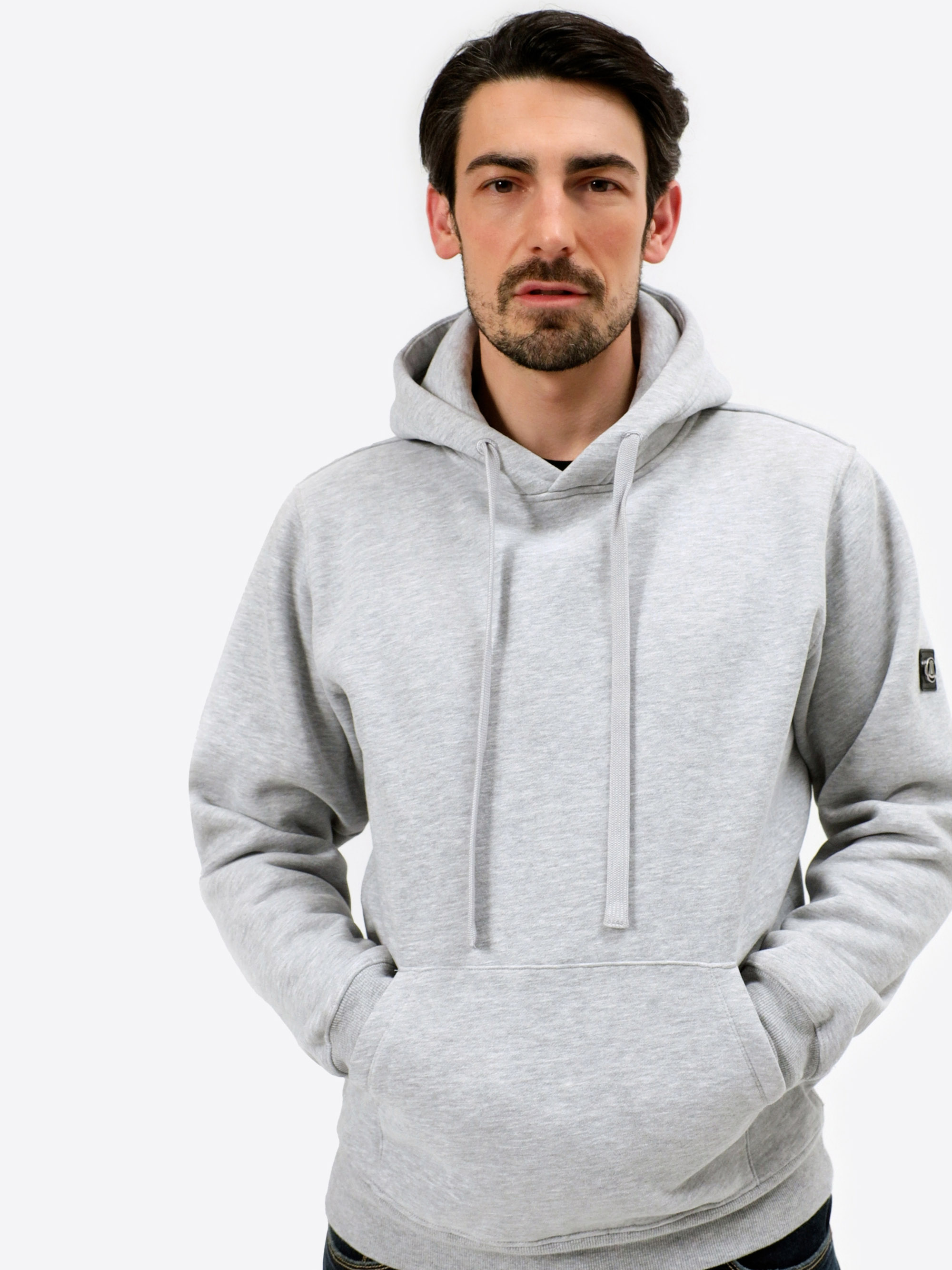 Ein Mann mit kurzen dunklen Haaren und einem Bart trägt einen hellgrauen Kapuzenpullover mit Kordelzug. Er hat die Hände in die Vordertasche gesteckt und steht vor einem schlichten weißen Hintergrund und schaut direkt in die Kamera.