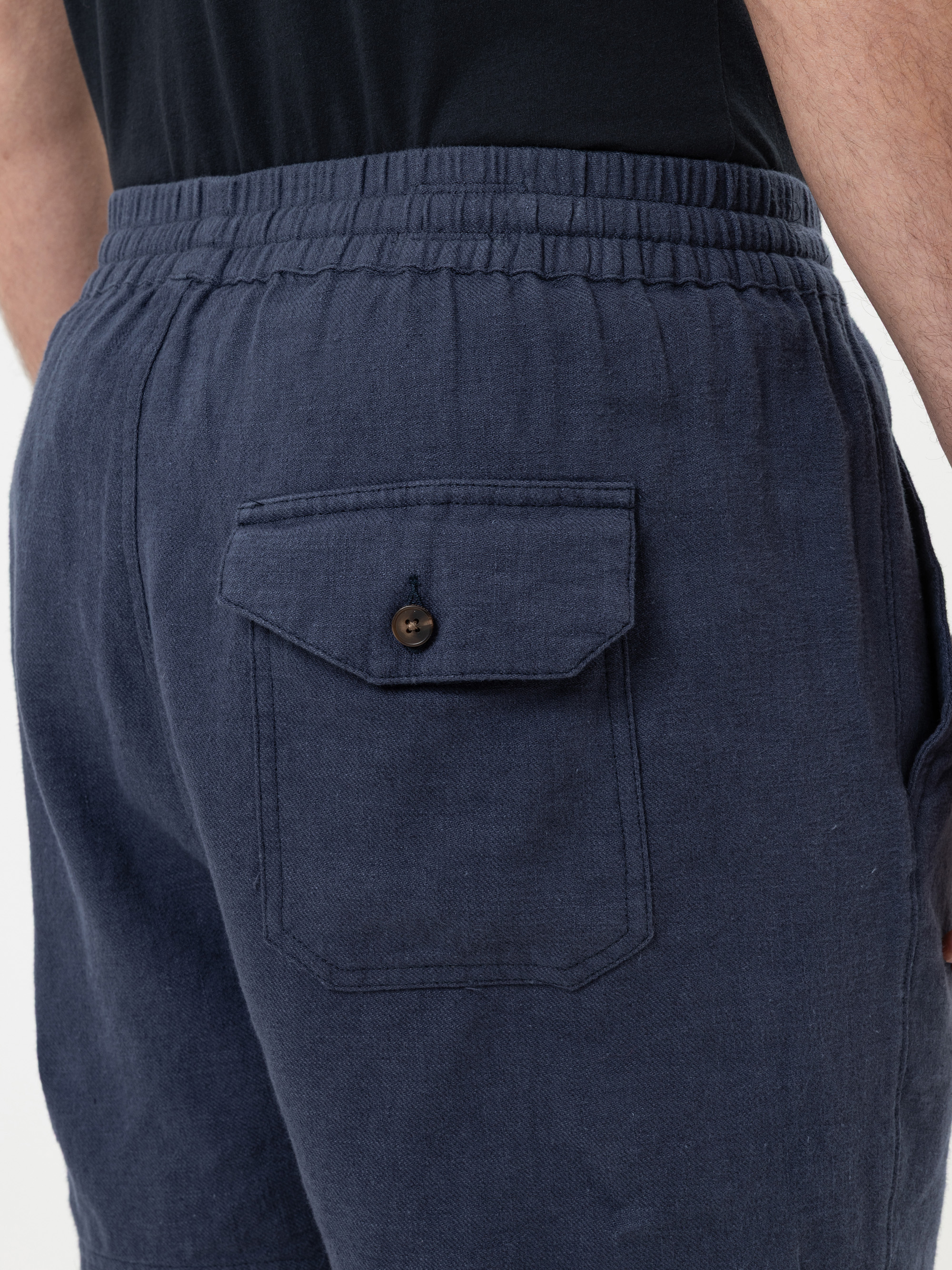 Nahaufnahme einer Person, die marineblaue Leinenshorts mit elastischem Bund und einer Gesäßtasche mit Klappe und braunem Knopf trägt. Die Person trägt außerdem ein schwarzes Hemd.
