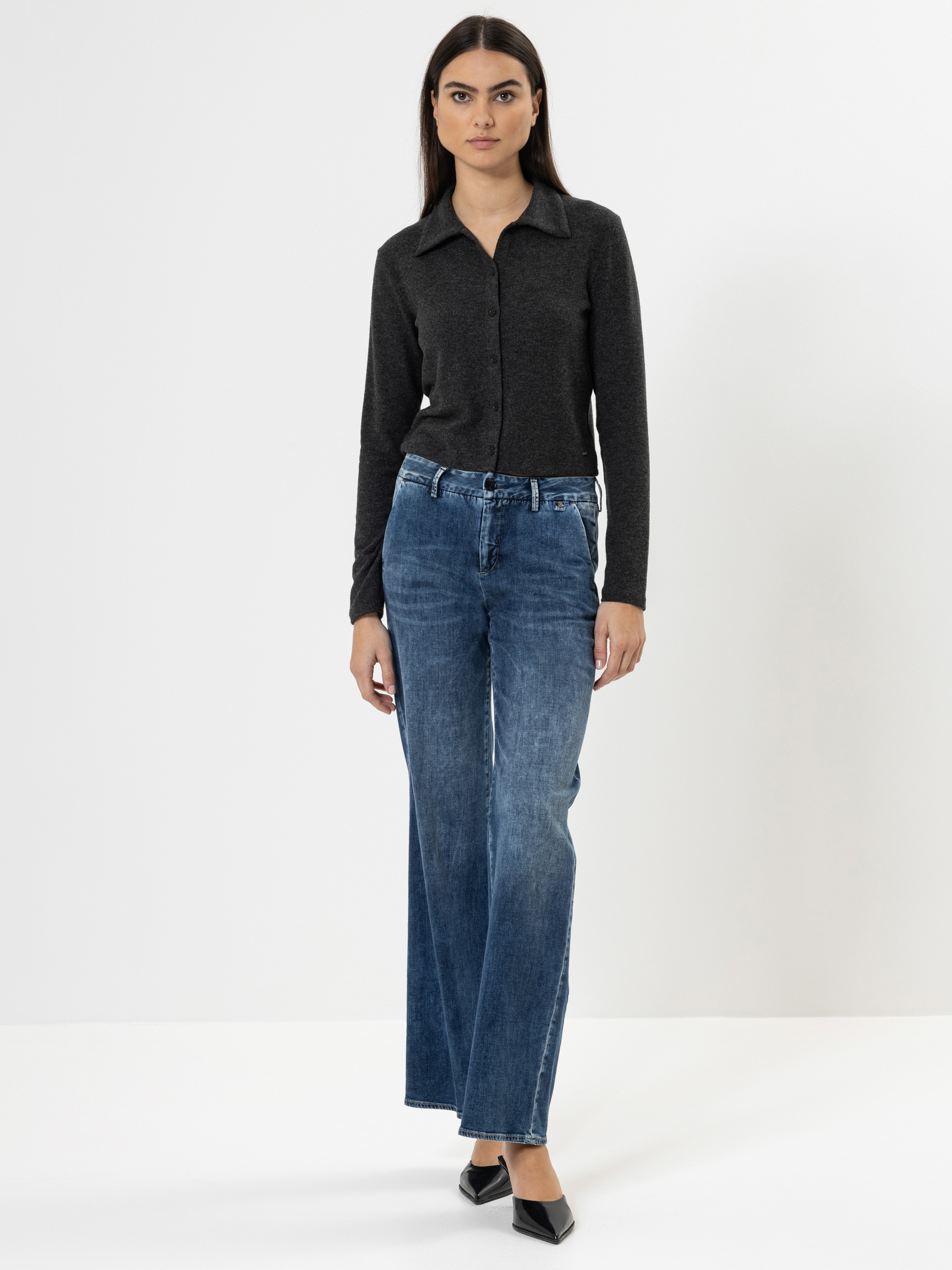 Eine Frau steht vor einem schlichten weißen Hintergrund und trägt ein dunkles langärmeliges Hemd mit Kragen, eine blaue Jeans mit hoher Taille und weitem Bein sowie schwarze Schuhe mit spitzen Zehen. Sie hat glattes dunkles Haar und einen neutralen Ges