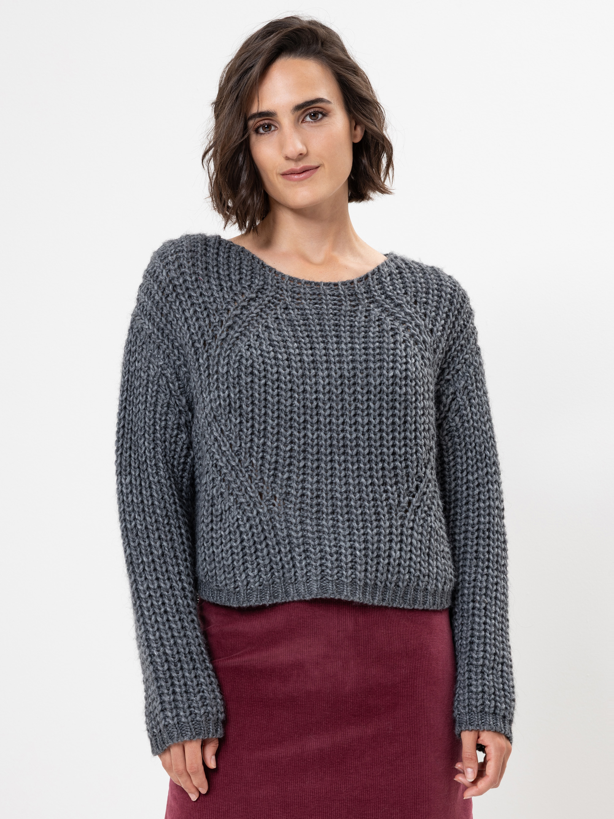 Eine Frau mit kurzen braunen Haaren, die einen dicken grauen Strickpullover und einen weinroten Rock trägt, steht vor einem schlichten weißen Hintergrund und blickt mit einem leichten Lächeln in die Kamera.