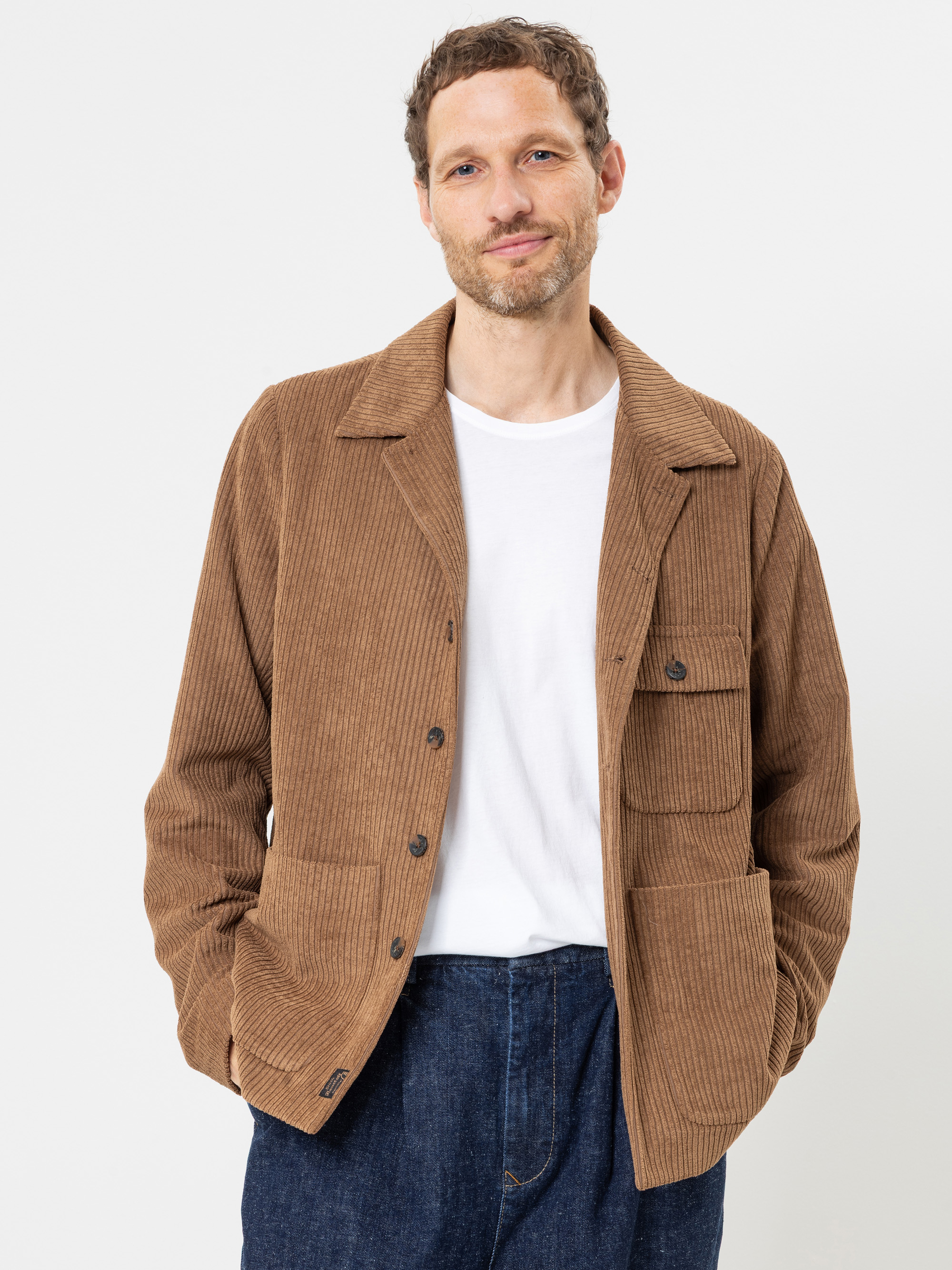 Ein Mann, der eine braune Cordjacke über einem weißen T-Shirt und blauen Jeans trägt, steht vor einem schlichten weißen Hintergrund, lächelt und hat die Hände in den Jackentaschen.