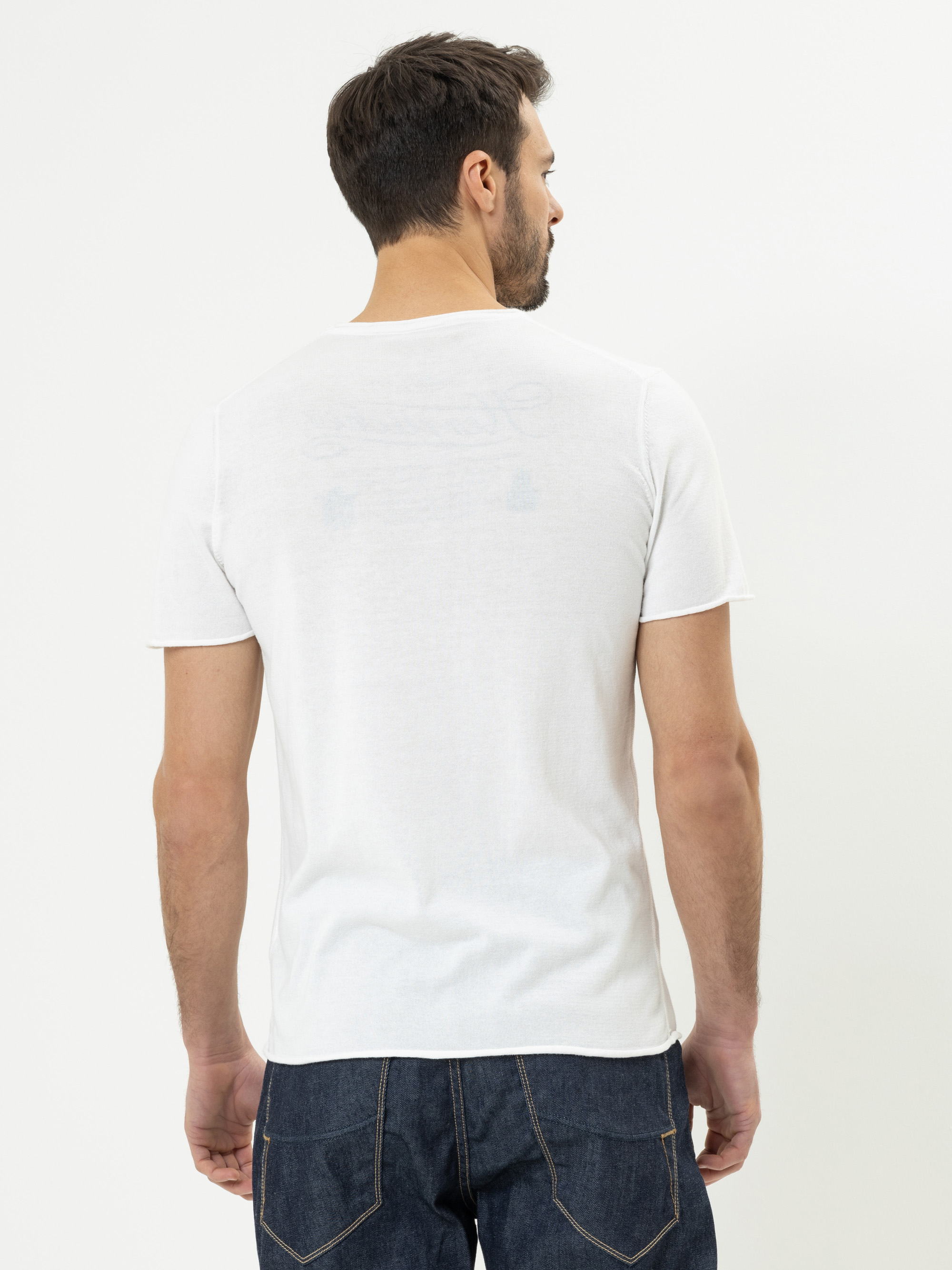 Ein Mann mit kurzen dunklen Haaren, Bart und Schnurrbart trägt ein schlichtes weißes T-Shirt und dunkelblaue Jeans. Er steht mit dem Gesicht von der Kamera weg vor einem weißen Hintergrund.