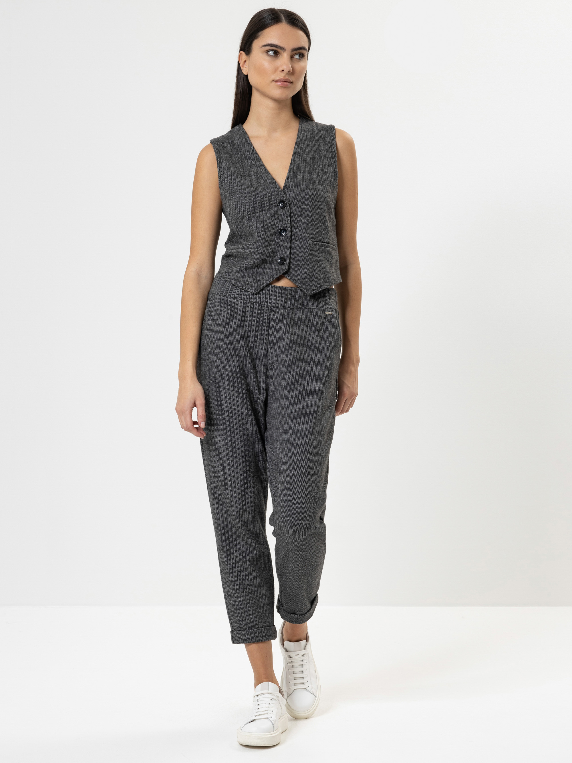 Eine Frau steht vor einem schlichten weißen Hintergrund. Sie trägt eine ärmellose graue Weste mit Knöpfen und eine dazu passende graue Hose, gepaart mit weißen Turnschuhen. Sie hat langes glattes dunkles Haar und einen neutralen Gesichtsausdruck.