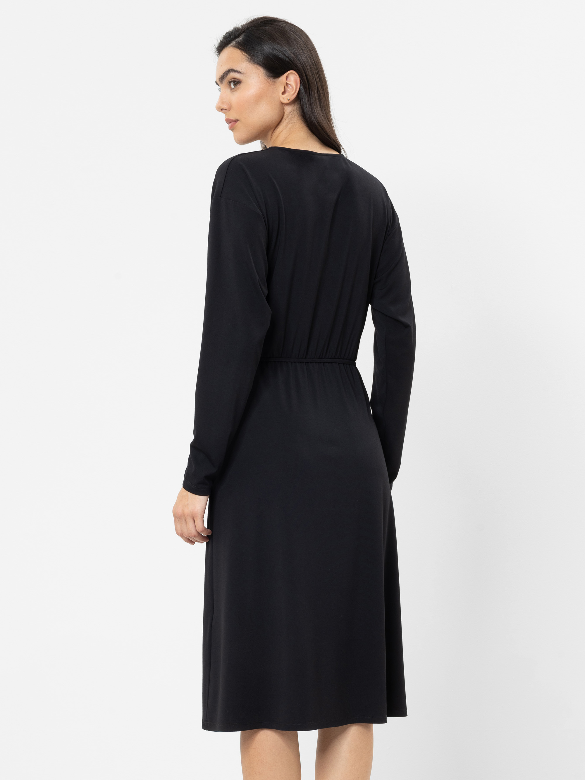 Eine Frau mit glattem, dunklem Haar trägt ein langärmeliges, knielanges schwarzes Kleid. Sie steht mit dem Rücken zur Kamera und blickt über die Schulter, vor einem schlichten weißen Hintergrund.