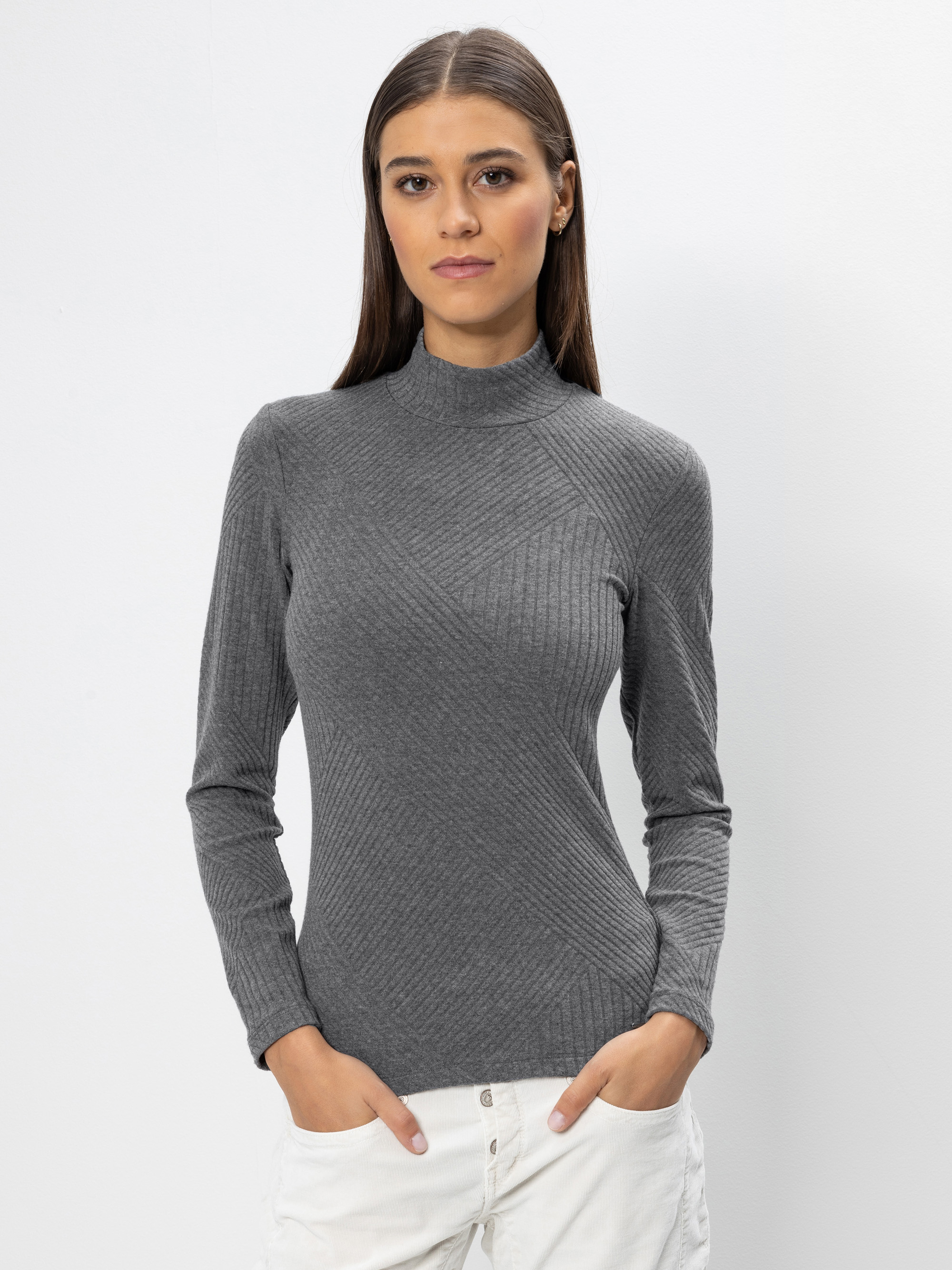 Eine Frau mit glattem braunem Haar trägt einen grauen Rollkragenpullover mit Rippenmuster und eine weiße Hose. Sie steht vor einem schlichten weißen Hintergrund und blickt mit neutraler Miene und den Händen in den Taschen in die Kamera.
