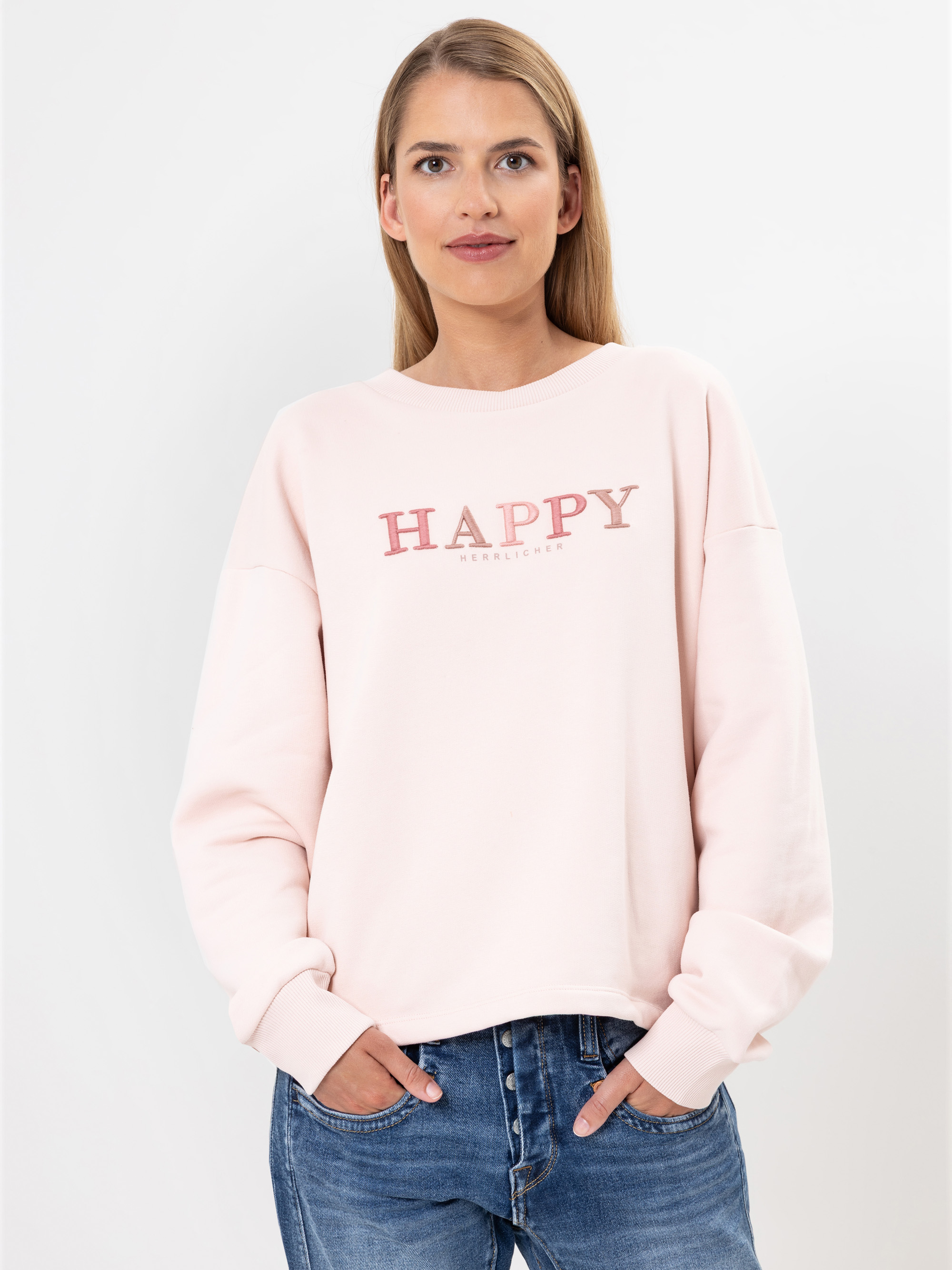 Eine Frau mit langen blonden Haaren trägt ein hellrosa Sweatshirt mit dem Aufdruck HAPPY und blaue Jeans. Sie steht vor einem schlichten weißen Hintergrund und hat die Hände in den Taschen.