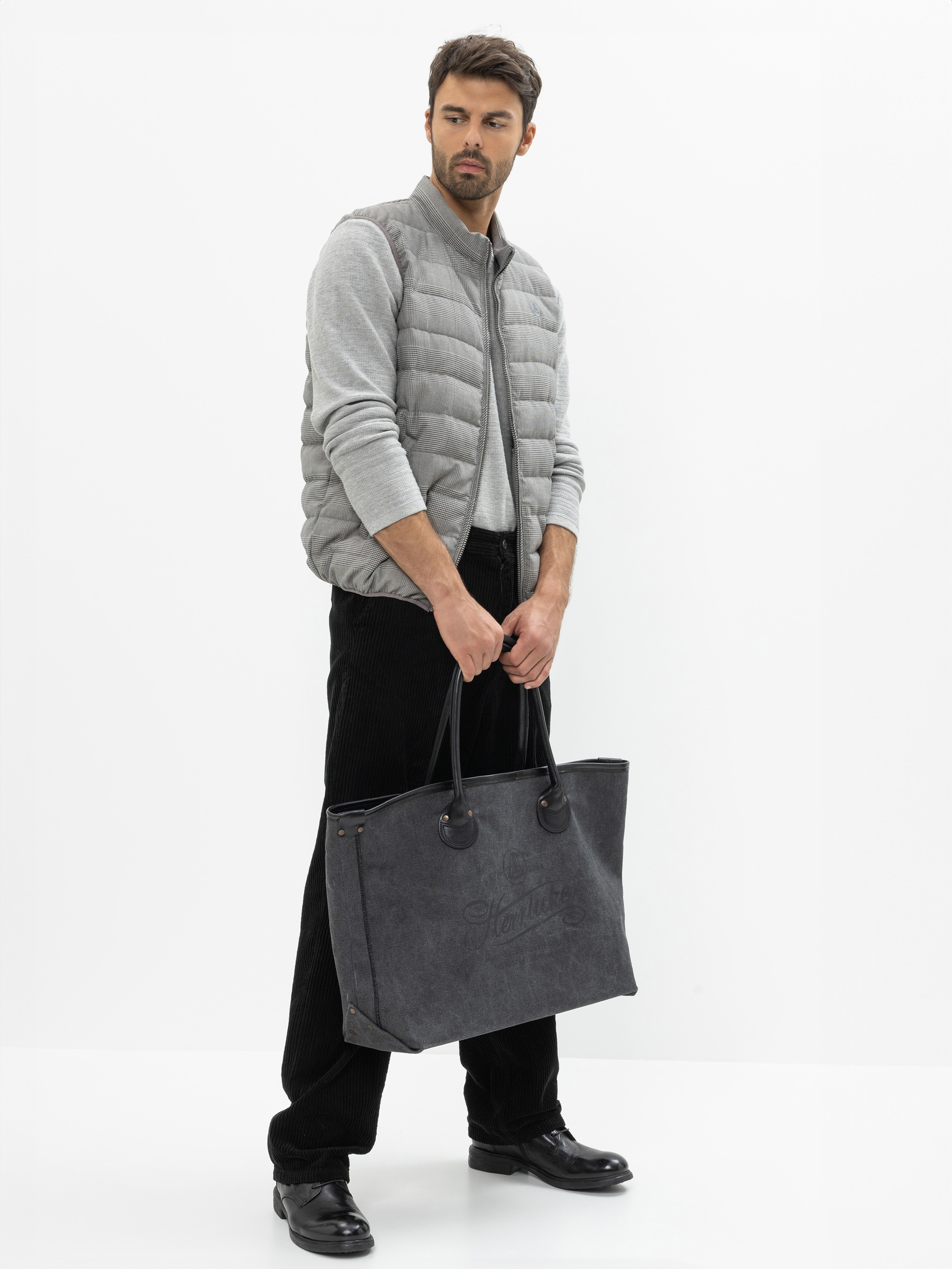 Ein Mann mit kurzen braunen Haaren und einem Bart trägt eine hellgraue Steppweste über einem grauen Pullover, schwarze Hosen und schwarze Schuhe und hält eine große dunkelgraue Tragetasche. Er steht vor einem schlichten weißen Hintergrund.