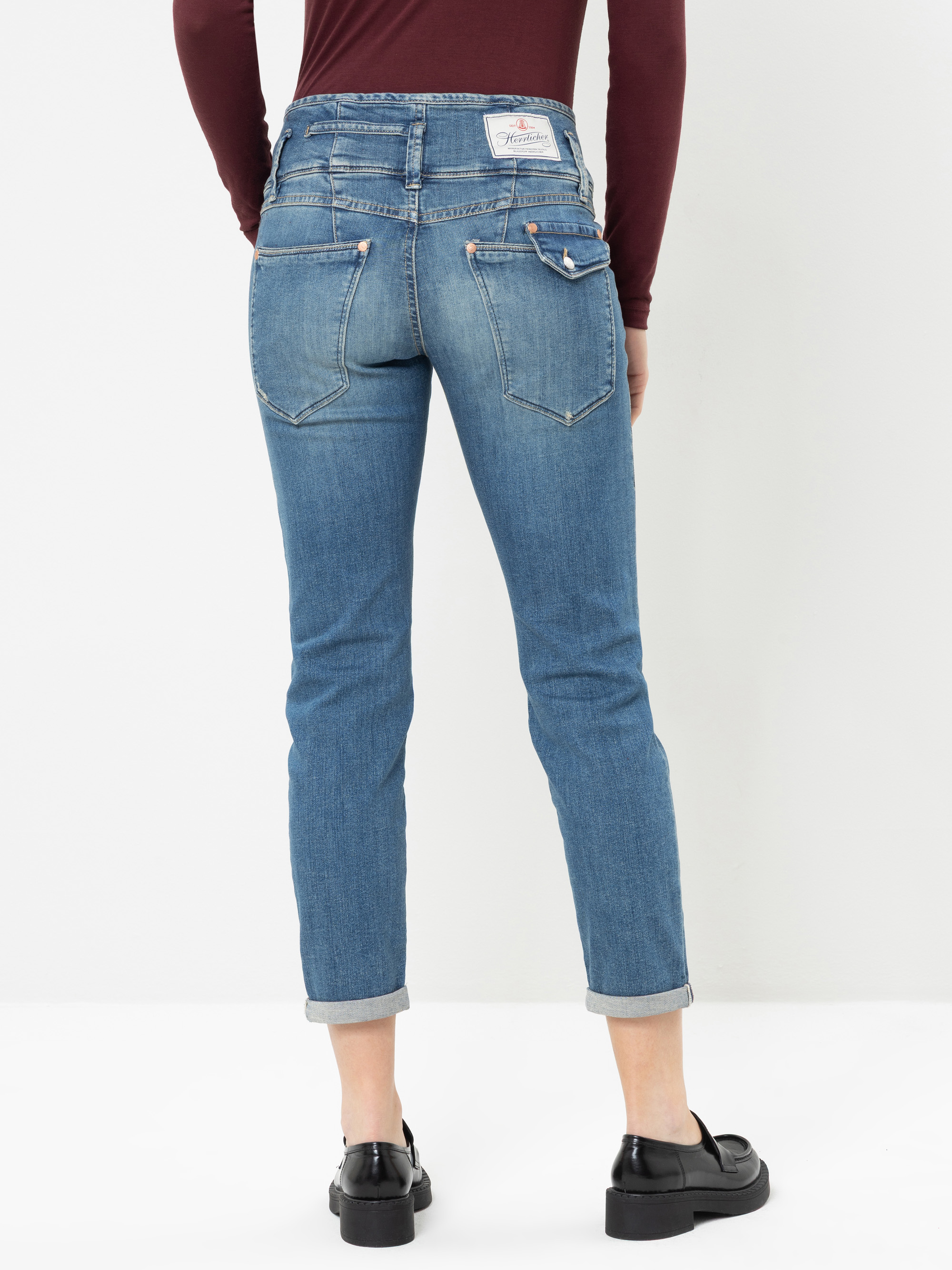 Eine Person, die hellblaue Jeans mit Umschlag, schwarze Schuhe und ein weinrotes Langarm-Oberteil trägt und mit dem Rücken zur Kamera vor einem schlichten weißen Hintergrund steht.