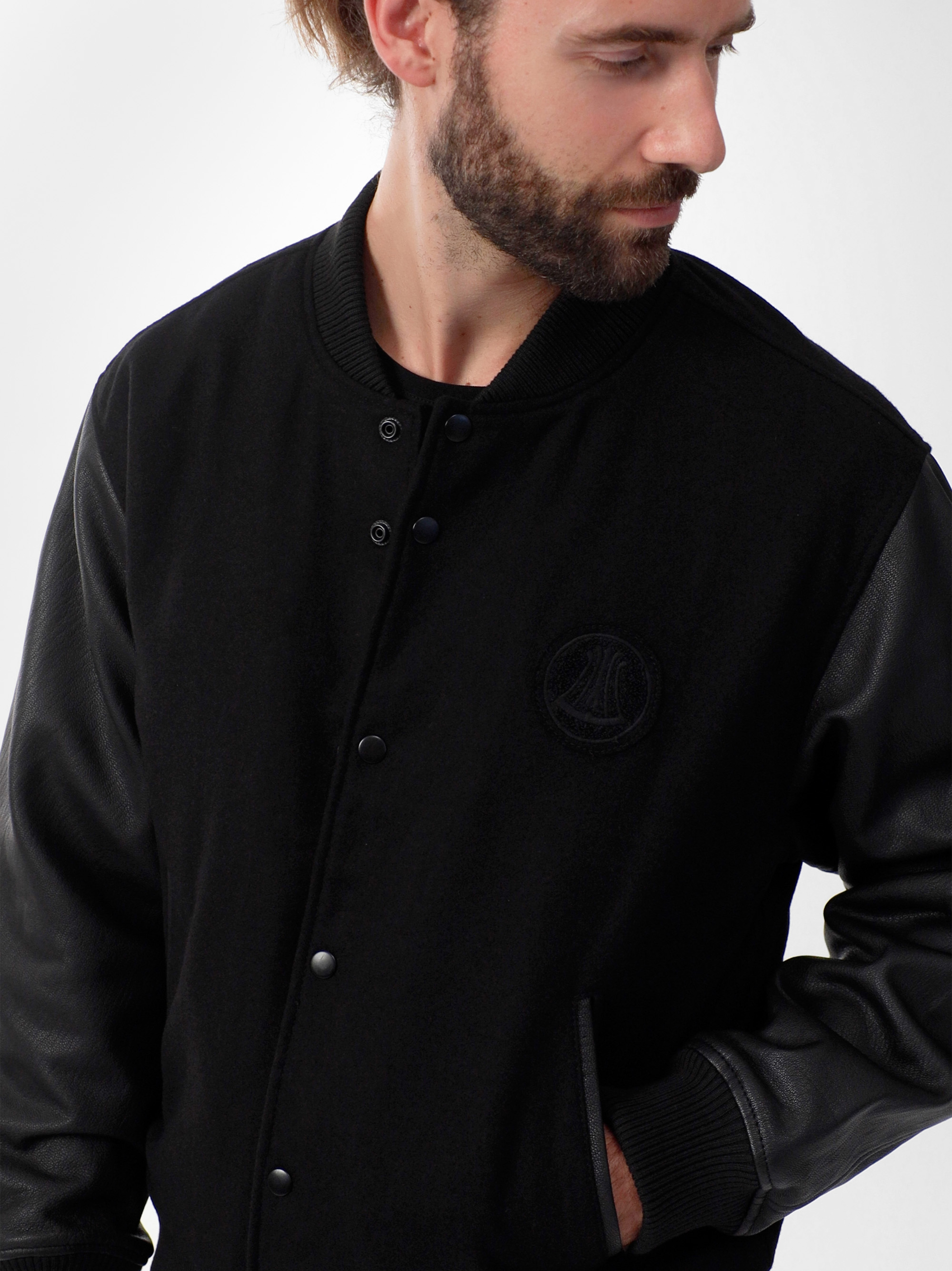 Ein bärtiger Mann trägt eine schwarze Sportjacke mit Lederärmeln und hat die linke Hand in der Hosentasche. Er blickt nach unten und zur Seite vor einem schlichten Hintergrund.