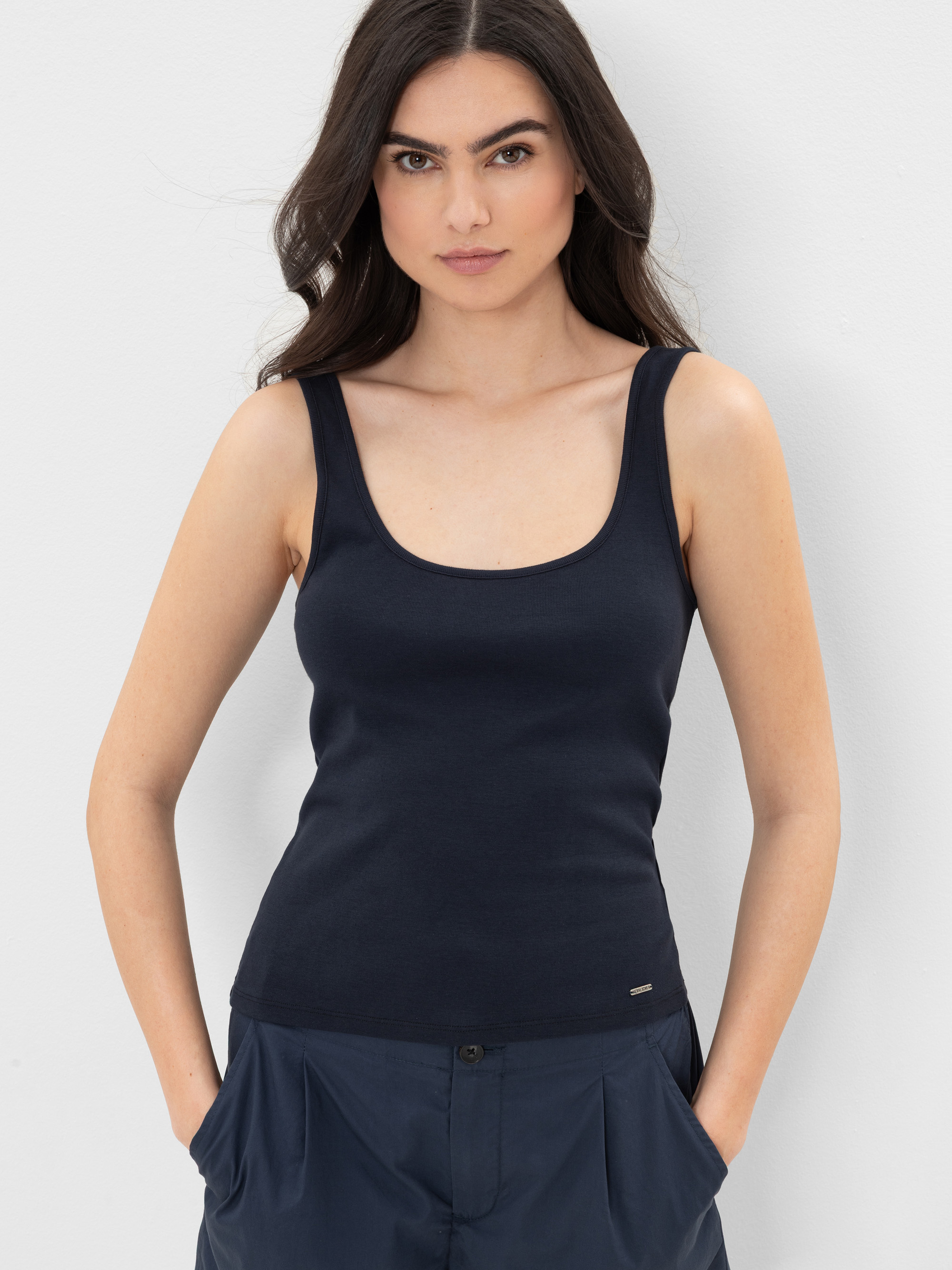Eine Frau mit langen dunklen Haaren trägt ein marineblaues ärmelloses Tank-Top und eine dazu passende marineblaue Hose. Sie steht vor einem schlichten weißen Hintergrund, hat die Hände in den Taschen und schaut direkt in die Kamera.