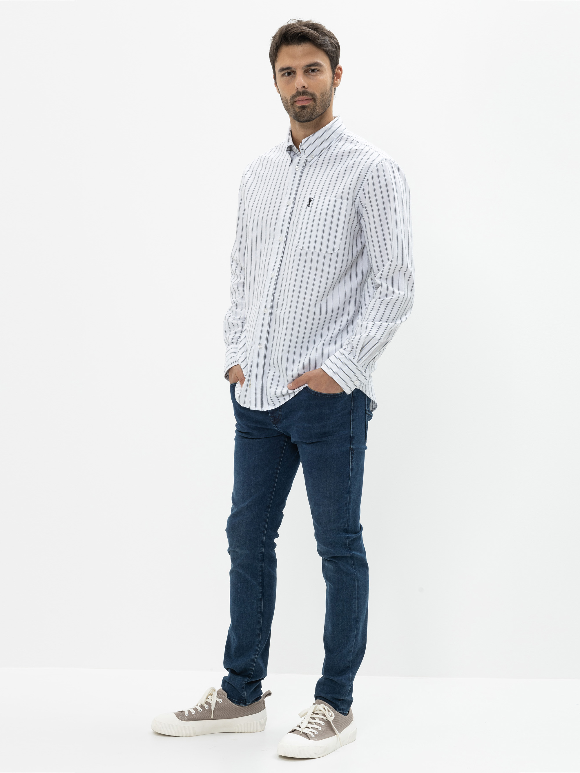 Ein Mann steht vor einem weißen Hintergrund und trägt ein weißes Button-up-Hemd mit vertikalen marineblauen Streifen, blaue Jeans und weiße Turnschuhe mit grauen Details. Er hat die Hände in den Taschen und einen neutralen Gesichtsausdruck.