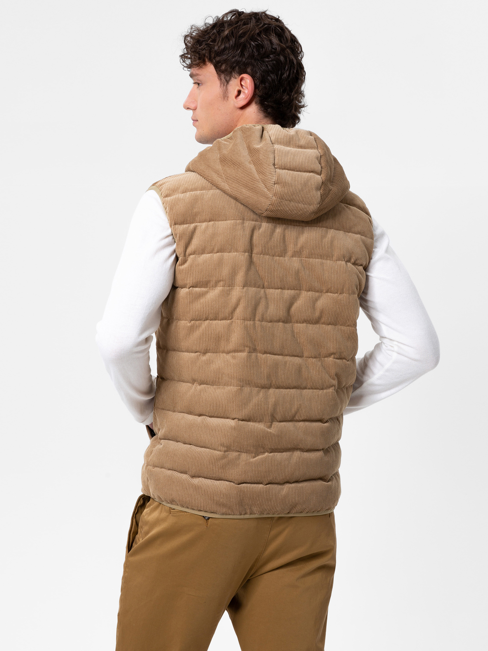 Ein Mann, der eine hellbraune gesteppte Kapuzenweste über einem weißen langärmeligen Hemd und einer braunen Hose trägt, steht mit dem Rücken zur Kamera vor einem schlichten weißen Hintergrund.