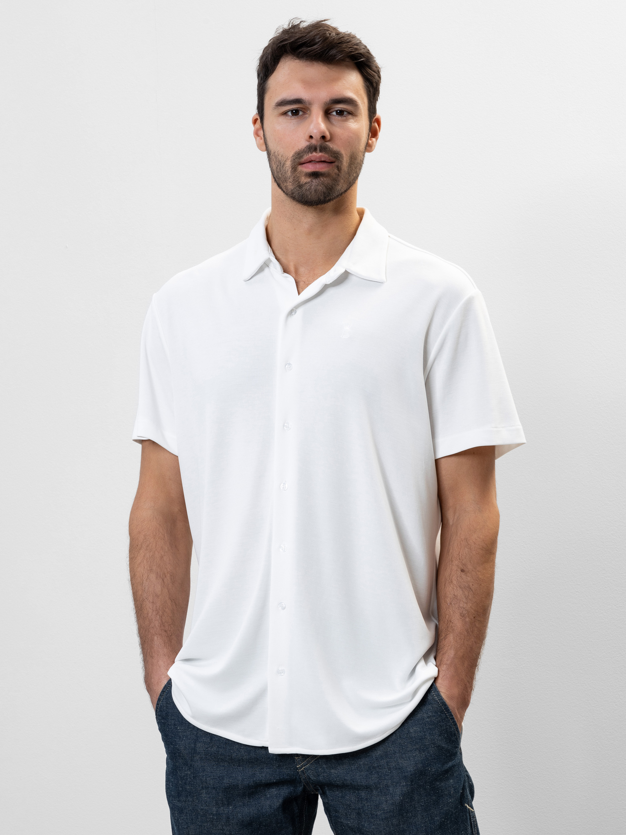 Ein Mann mit kurzen dunklen Haaren und Bart trägt ein weißes, kurzärmeliges Button-up-Hemd und eine dunkle Hose und steht mit den Händen in den Taschen vor einem schlichten weißen Hintergrund.