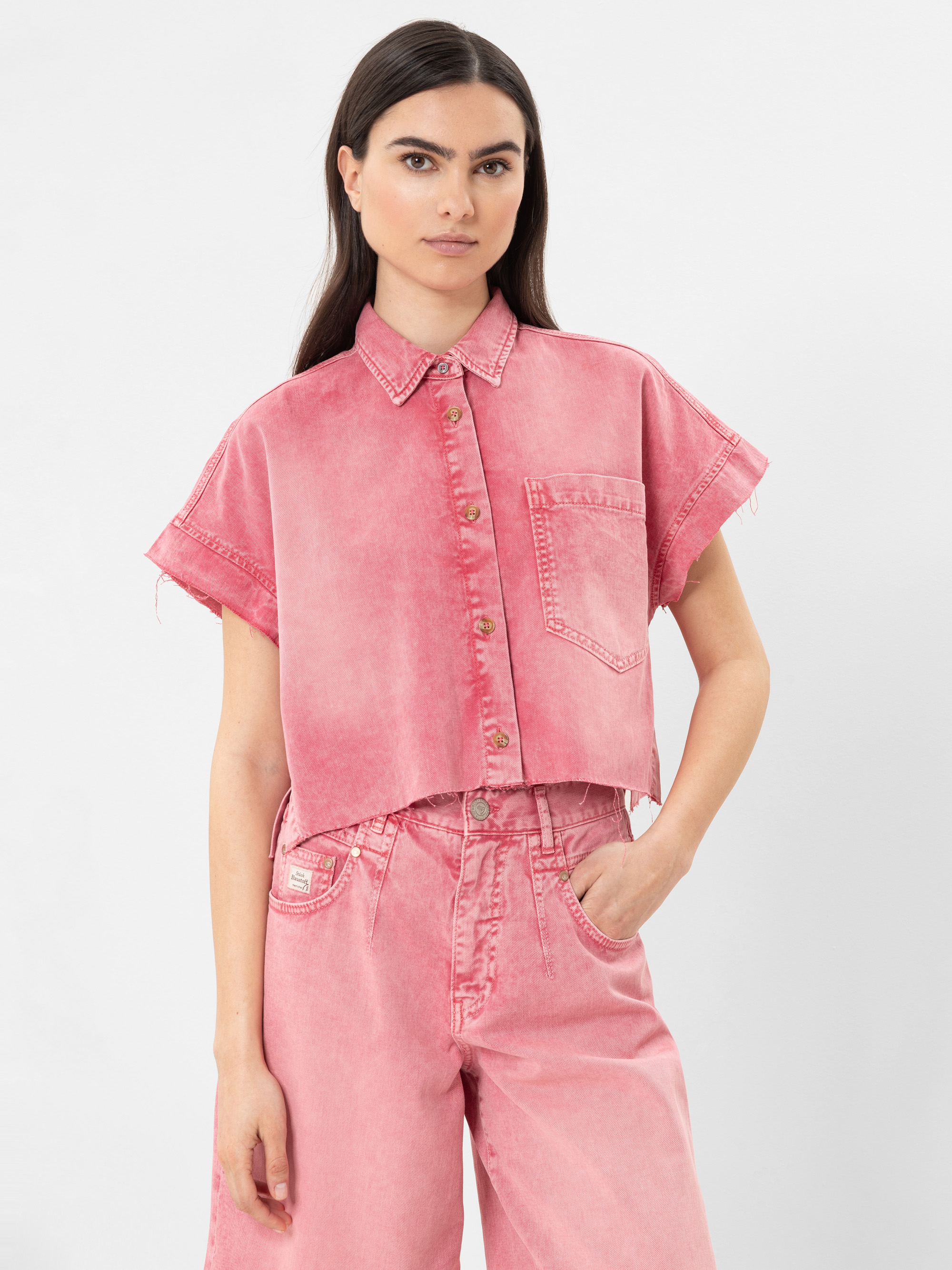 Eine Frau, die ein lockeres, kurzärmeliges, abgeschnittenes rosa Jeanshemd mit Kragen und Vordertasche trägt, gepaart mit einer passenden hochtaillierten rosa Jeans, steht vor einem schlichten weißen Hintergrund.