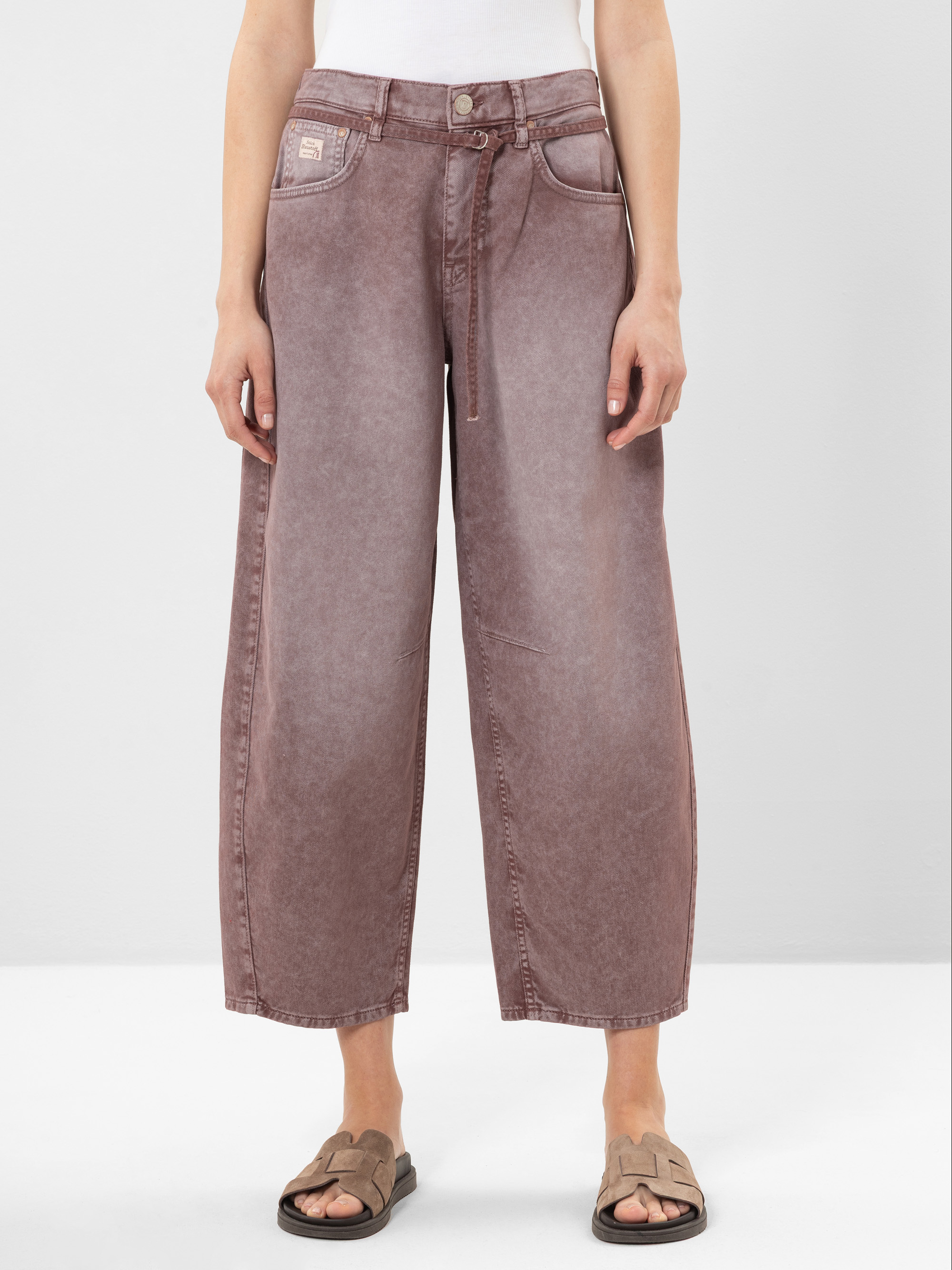 Eine Person, die eine verblichene lilafarbene Jeans mit weitem Bein und hoher Taille sowie einen Gürtel zum Binden auf der Vorderseite, ein weißes Oberteil und beigefarbene Slippersandalen trägt, steht vor einem schlichten hellen Hintergrund.