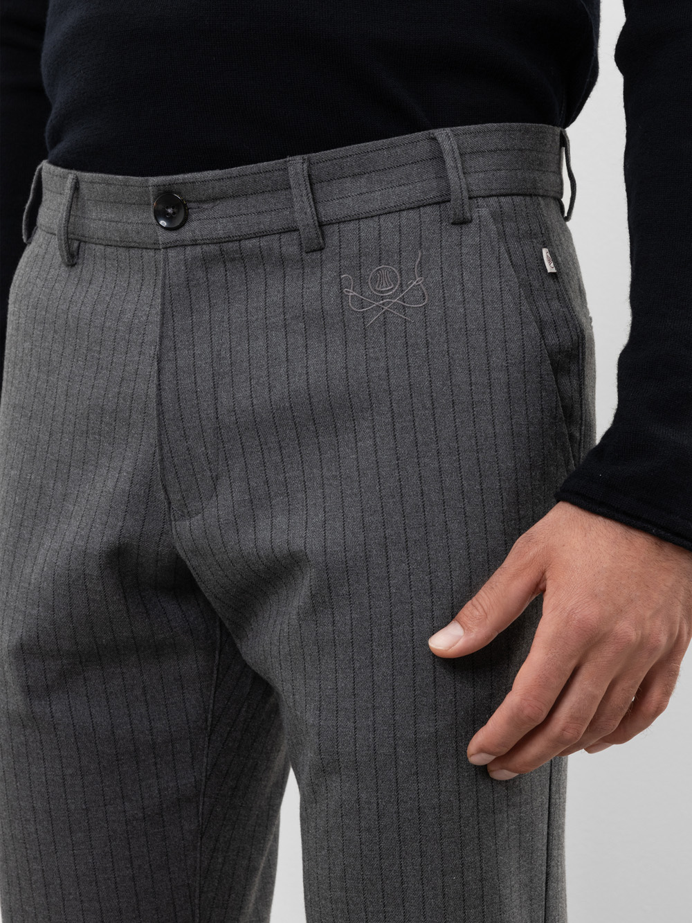 Eine Person trägt eine graue Nadelstreifenhose mit einem kleinen gestickten Tennisschläger- und Ballmuster in der Nähe der Tasche. Die rechte Hand der Person ist sichtbar, und sie trägt ein schwarzes langärmeliges Hemd.
