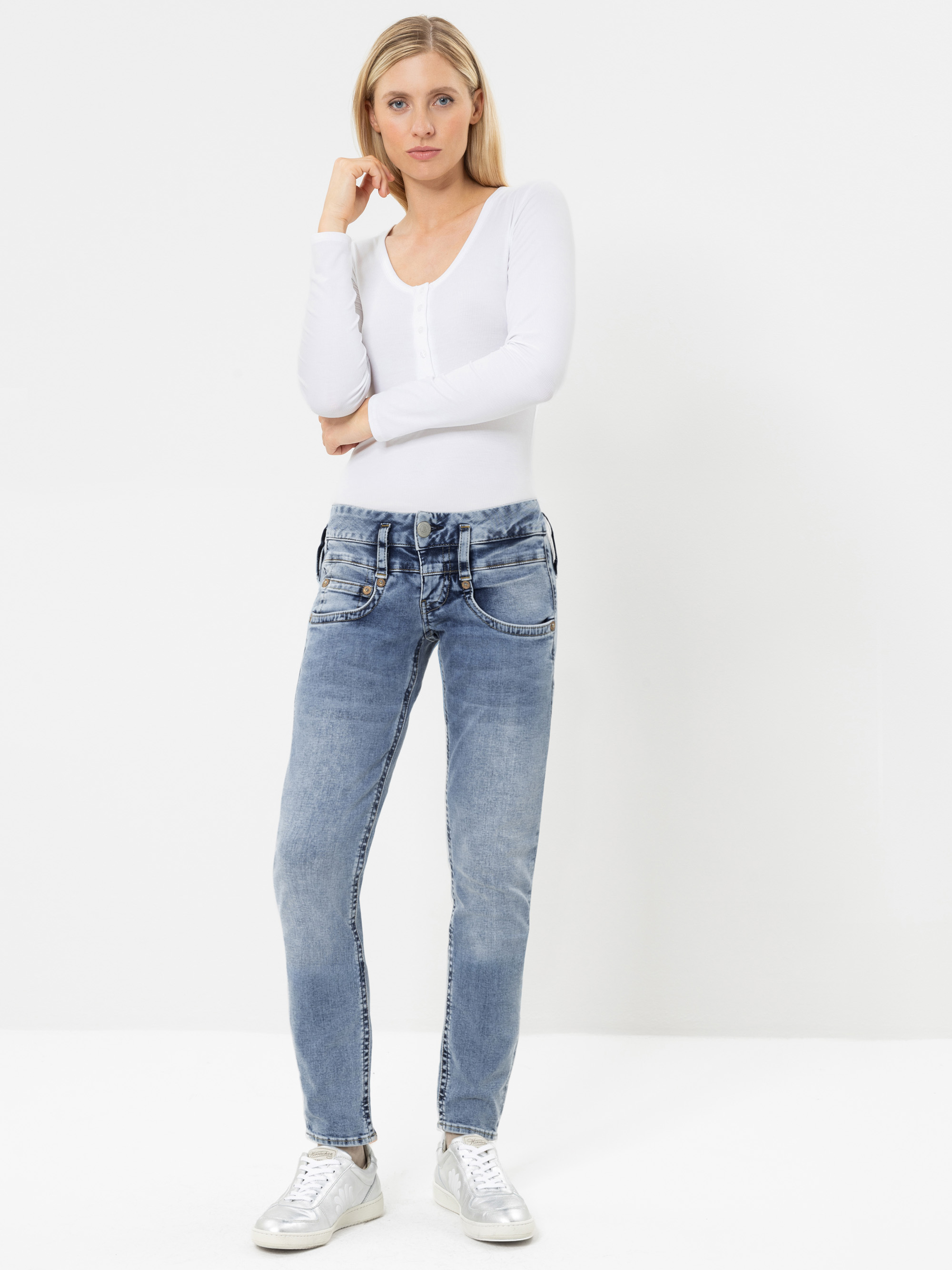 Eine Frau, die ein weißes Langarm-Oberteil, hellblaue Jeans und weiße Sneaker trägt, steht vor einem schlichten weißen Hintergrund und blickt in die Kamera, wobei sie eine Hand nahe am Kinn hält.