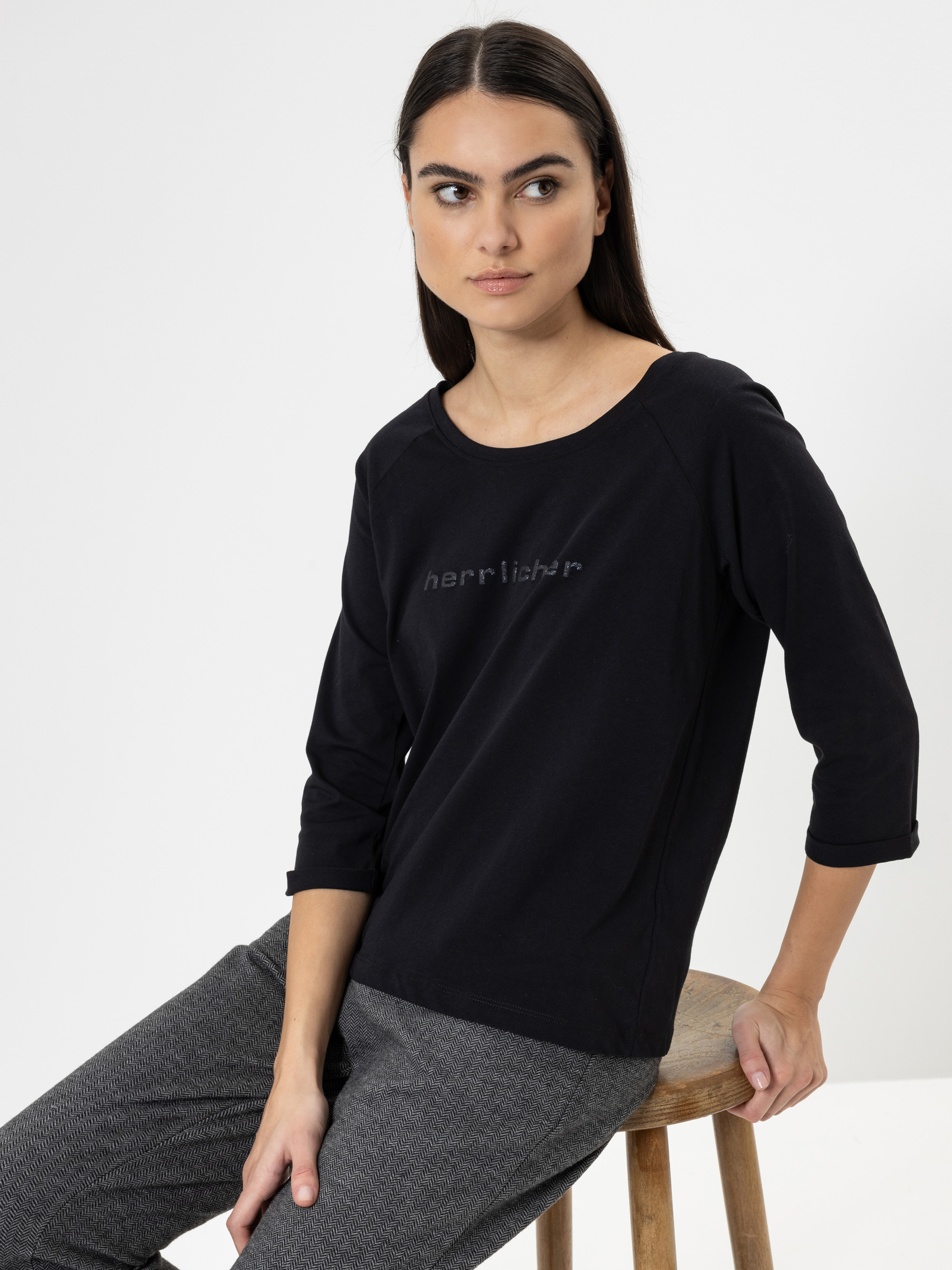 Eine Frau mit langen dunklen Haaren, die ein schwarzes Oberteil mit der Aufschrift herrlicher und eine graue Hose trägt, sitzt auf einem Holzschemel vor einem schlichten weißen Hintergrund und blickt leicht nach links.