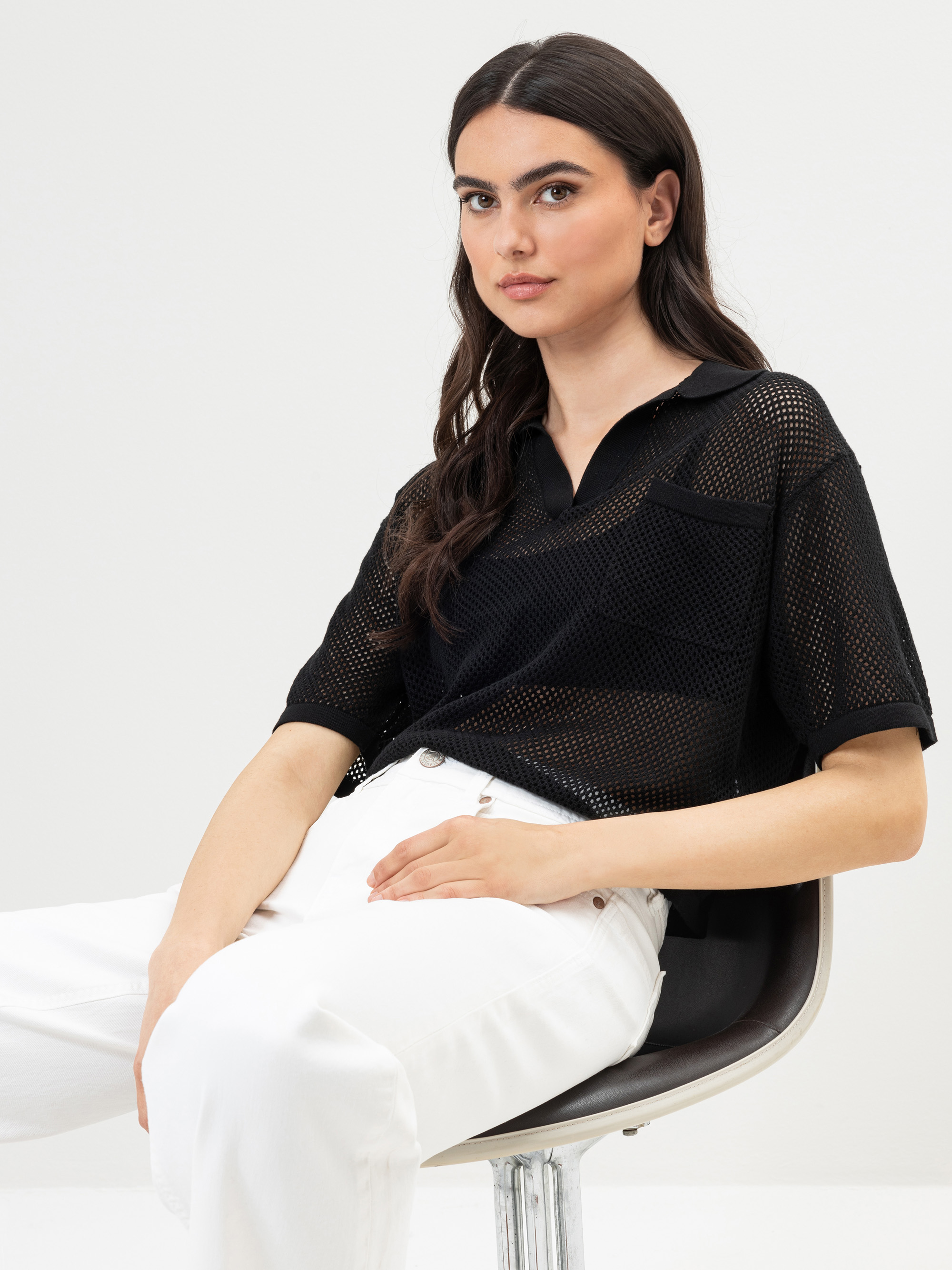 Eine Frau mit langen dunklen Haaren sitzt auf einem Stuhl und trägt ein schwarzes kurzärmeliges Netzoberteil mit Kragen und eine weiße Hose. Sie blickt leicht zur Seite und hat einen neutralen Gesichtsausdruck vor einem schlichten hellen Hintergrund.