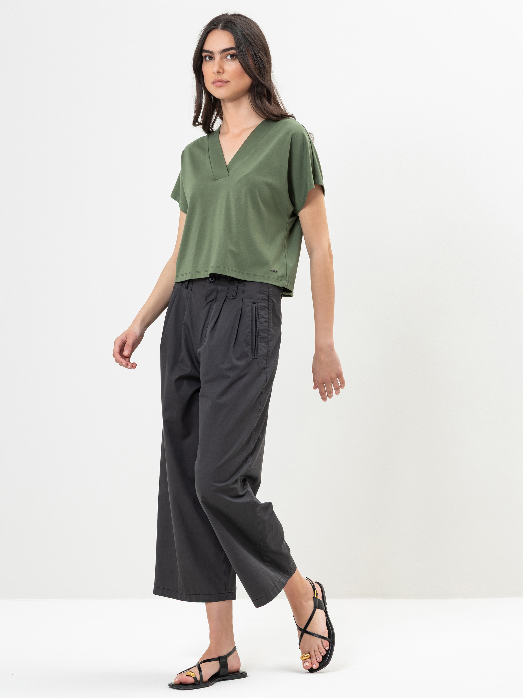 Eine Frau in einem lockeren, kurzärmeligen, olivgrünen Oberteil mit V-Ausschnitt und einer dunkelgrauen Hose mit weitem Bein steht vor einem schlichten weißen Hintergrund und trägt schwarze Sandalen.