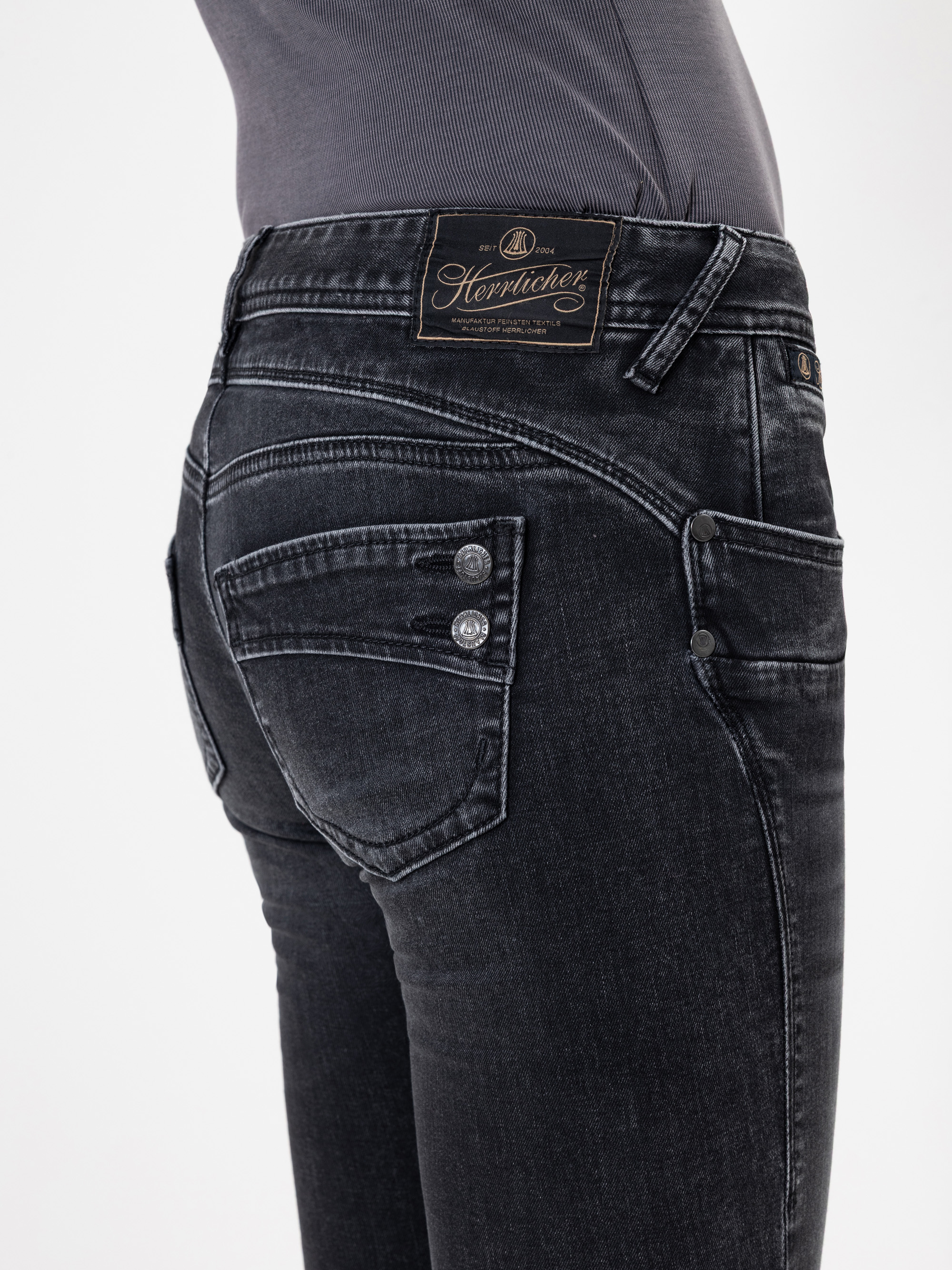 Nahaufnahme einer Person, die eine dunkel gewaschene Jeans mit Zierknöpfen an den Gesäßtaschen und einem schwarzen Herpatter-Etikett am Bund trägt. Die Person trägt außerdem ein tailliertes, dunkelgraues Oberteil.