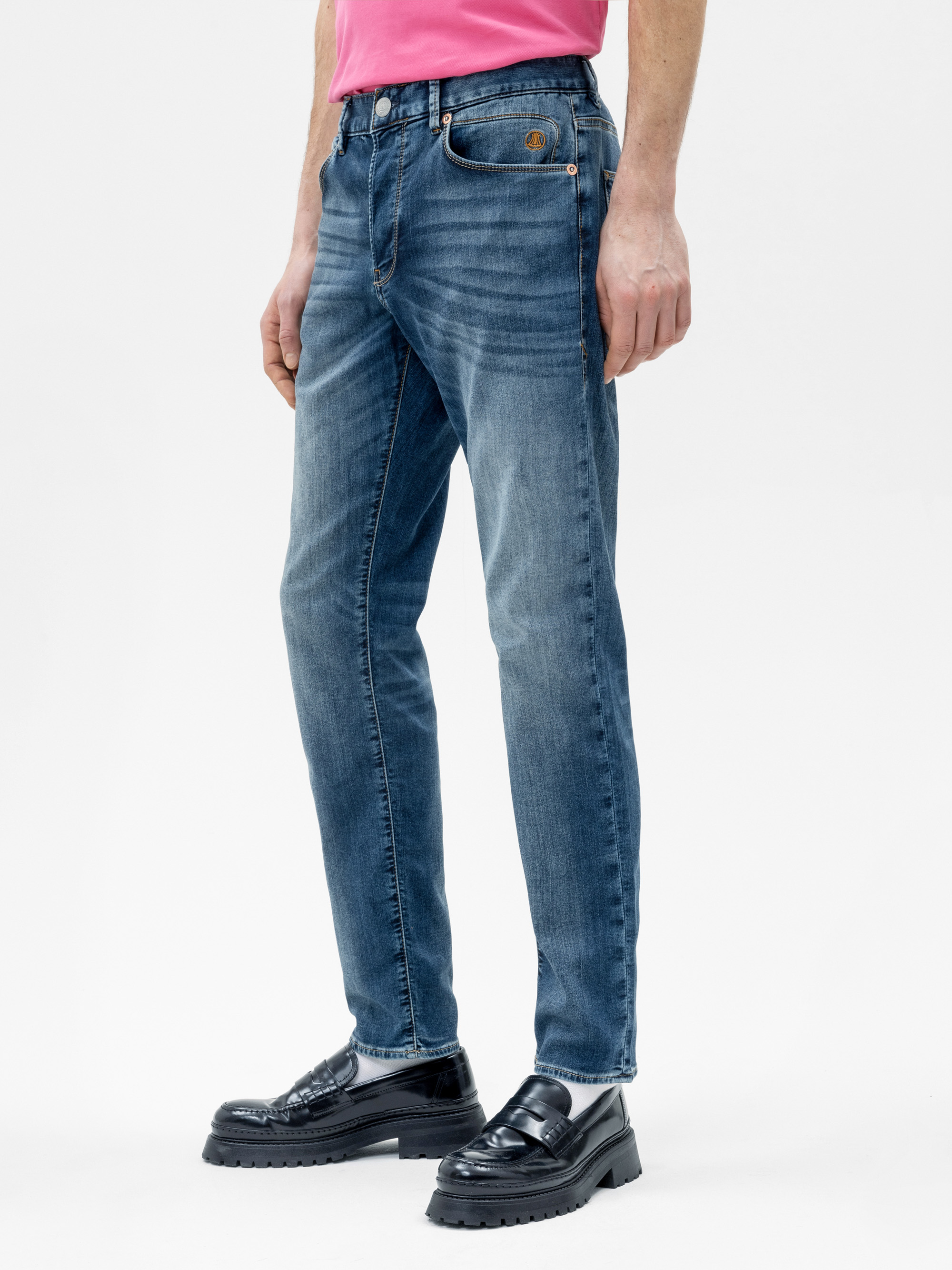 Eine Person, die ein rosa Hemd, eine blaue Slim-Fit-Jeans, weiße Socken und schwarze Halbschuhe trägt, steht vor einem schlichten weißen Hintergrund. Das Foto zeigt die Jeans und die untere Hälfte des Outfits.