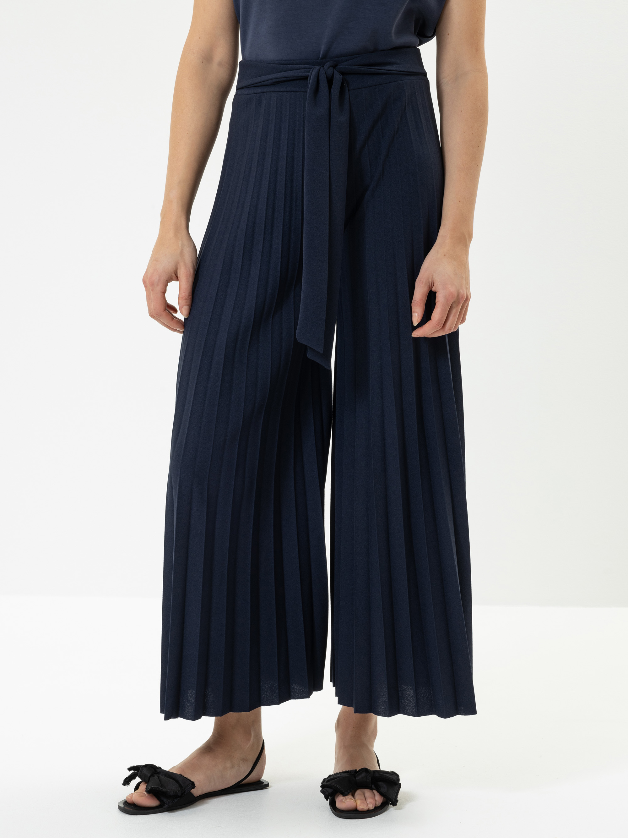 Eine Person, die eine marineblaue, hochtaillierte Hose mit weitem Bein und einem passenden Stoffgürtel trägt, dazu ein dunkles Oberteil und schwarze Slide-Sandalen mit Schleifen, steht vor einem schlichten hellen Hintergrund.