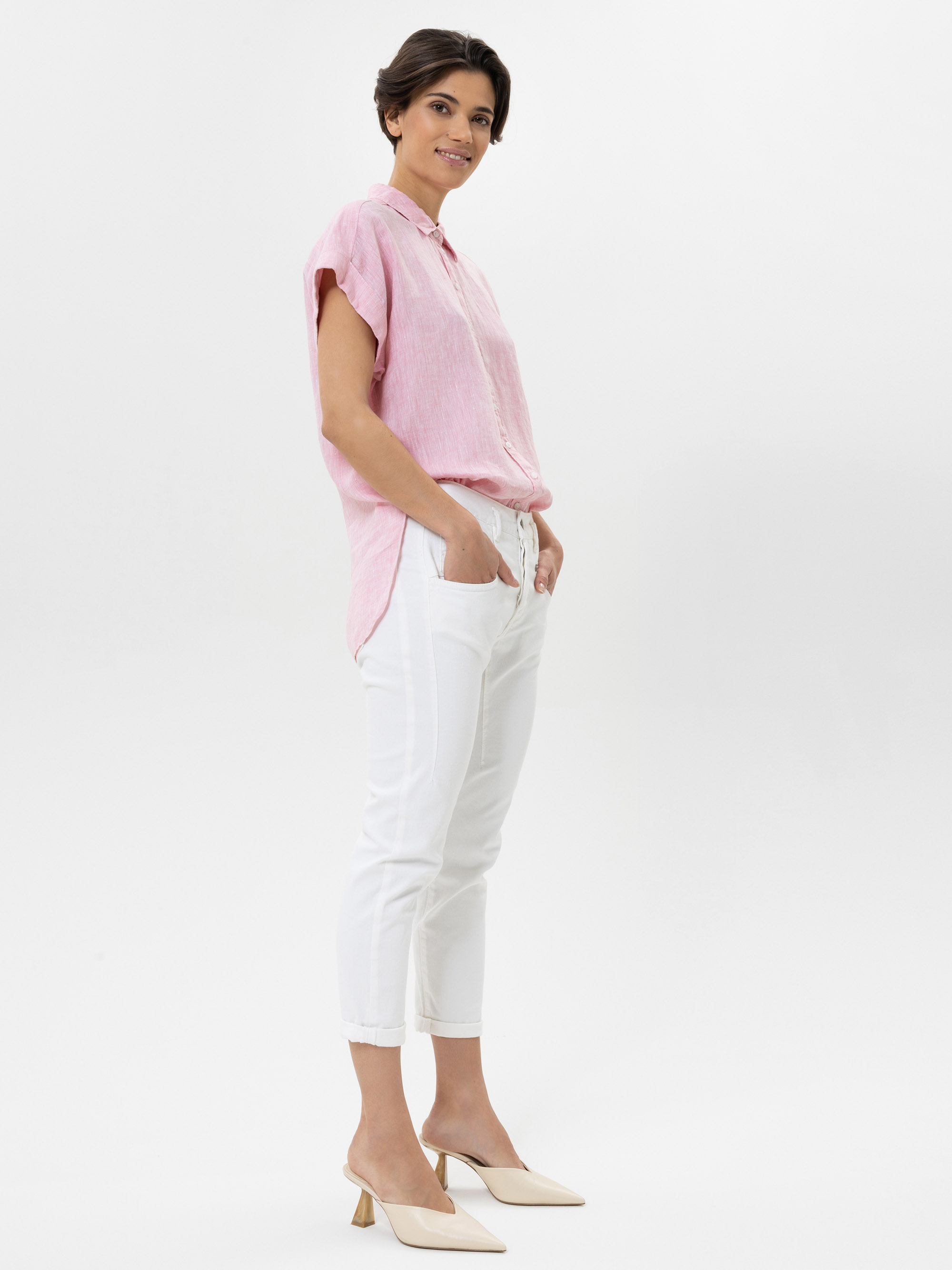 Eine Frau steht seitlich und lächelt. Sie trägt ein hellrosa kurzärmeliges Button-up-Hemd, eine weiße Hose und beigefarbene Schuhe mit Absatz vor einem schlichten weißen Hintergrund.