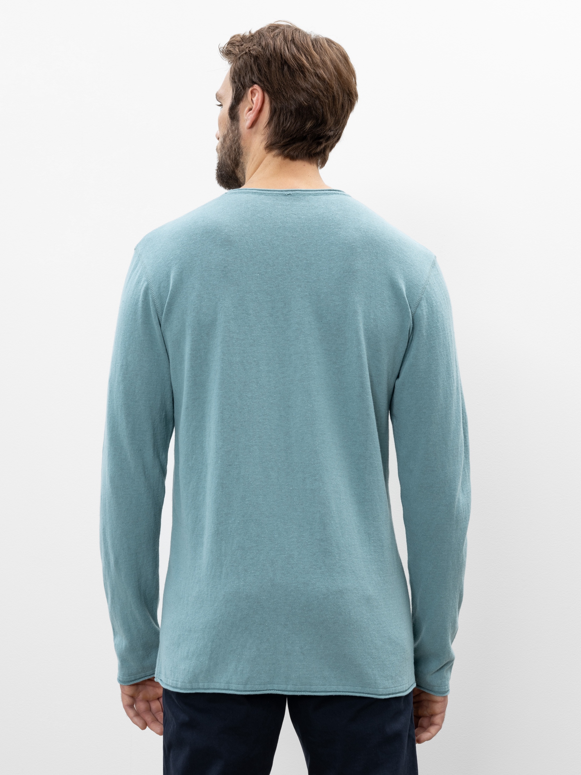 Ein Mann mit braunen Haaren, der ein hellblaues langärmeliges Hemd und eine dunkle Hose trägt, steht mit dem Gesicht zur Kamera vor einem schlichten weißen Hintergrund.