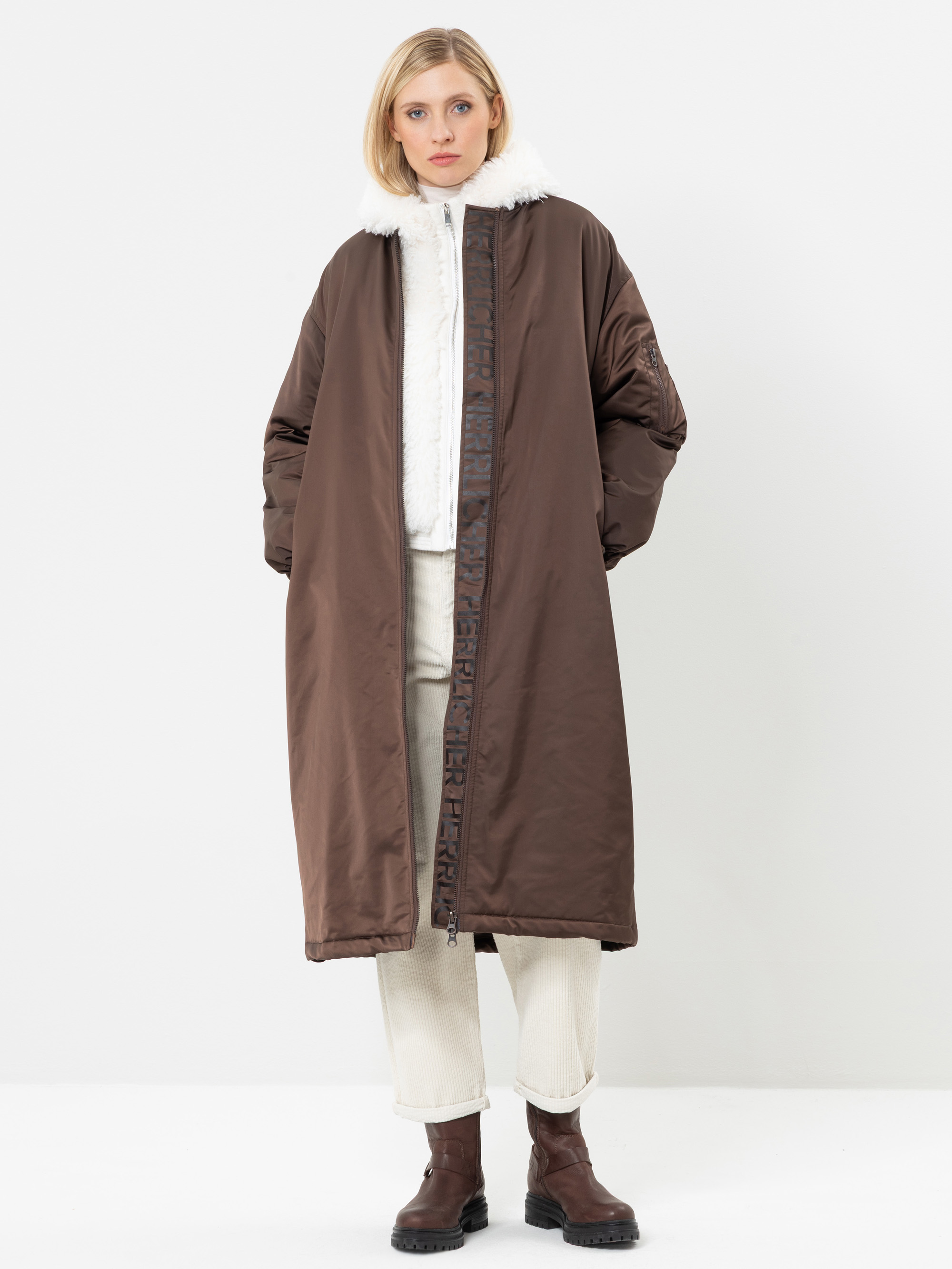 Eine Frau mit blonden Haaren trägt einen langen braunen Mantel mit einem weißen Pelzkragen, eine weiße Hose und braune Stiefel. Sie steht vor einem schlichten weißen Hintergrund und hat die Hände in den Manteltaschen.