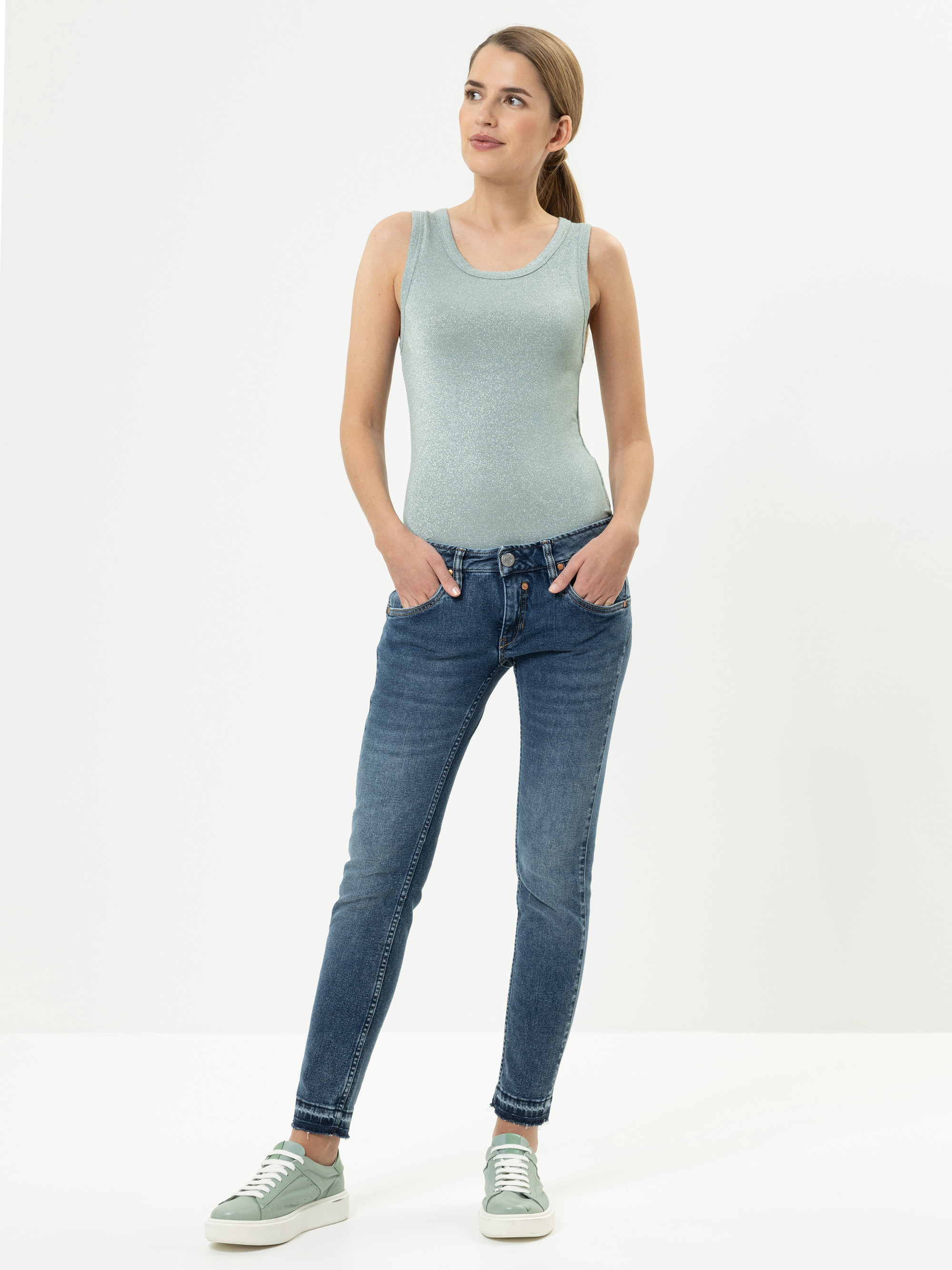 Eine Frau steht vor einem schlichten weißen Hintergrund und trägt ein hellblaues ärmelloses Oberteil, blaue Jeans und hellgrüne Turnschuhe. Sie hat die Hände in den Hosentaschen und blickt leicht zur Seite, ihr Haar ist zurückgebunden.