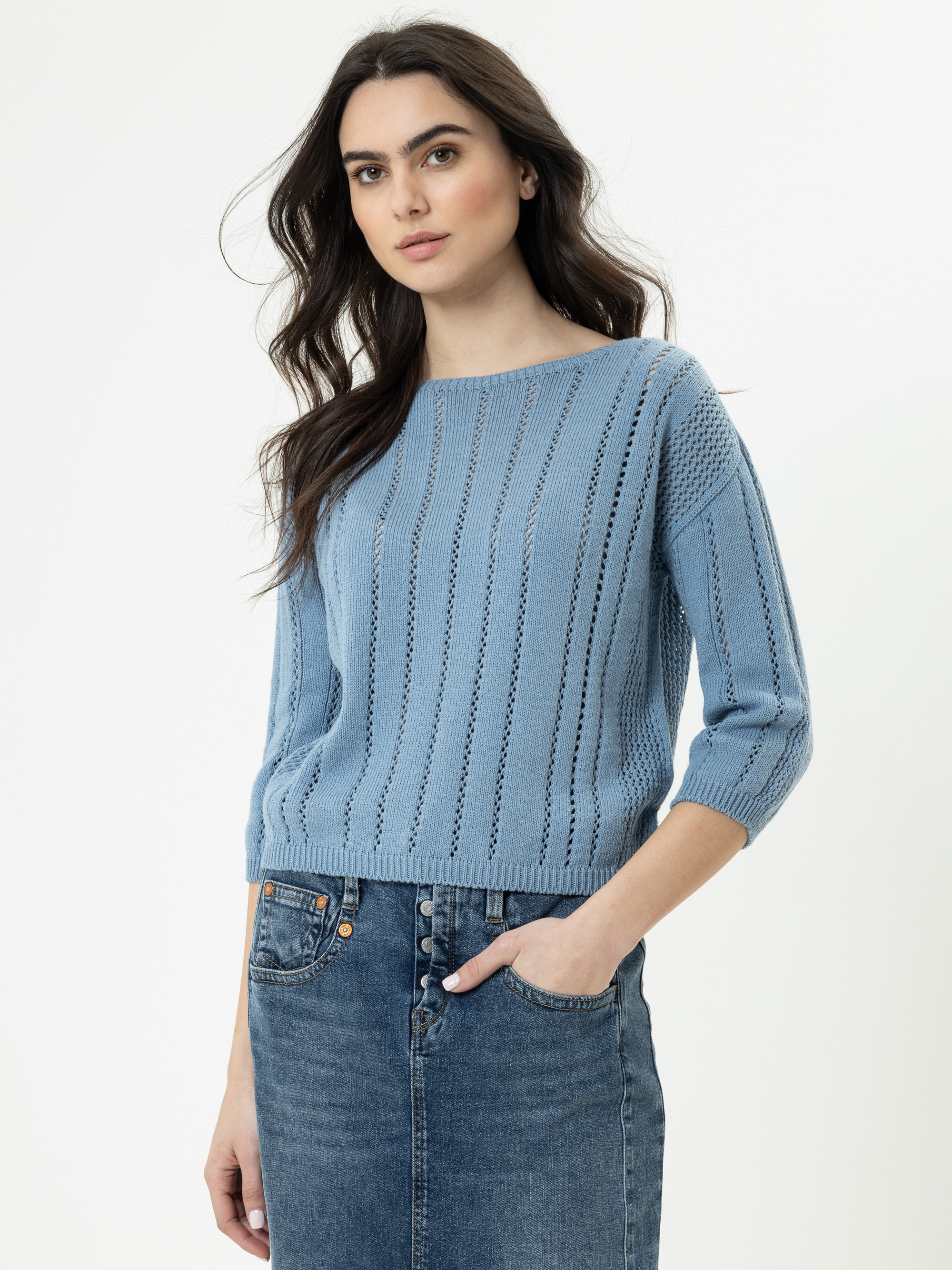 Eine Frau mit langen dunklen Haaren, die einen hellblauen Strickpullover und blaue Jeans trägt, steht vor einem schlichten weißen Hintergrund, hat eine Hand in der Tasche und einen neutralen Gesichtsausdruck.