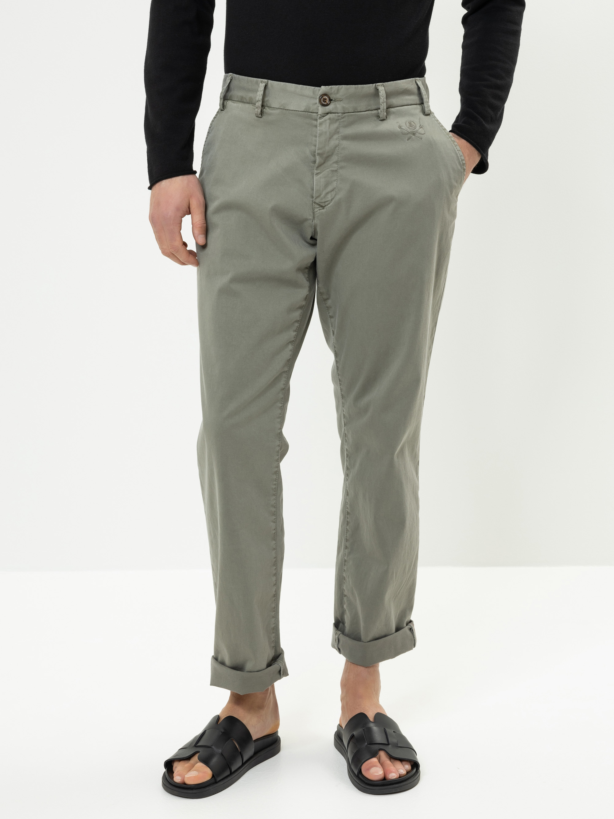 Eine Person, die eine aufgerollte olivgrüne Hose, ein schwarzes langärmeliges Oberteil und schwarze Sandalen trägt, steht vor einem schlichten weißen Hintergrund. Ihre Hände befinden sich in den Taschen, und ihr Gesicht ist nicht zu sehen.