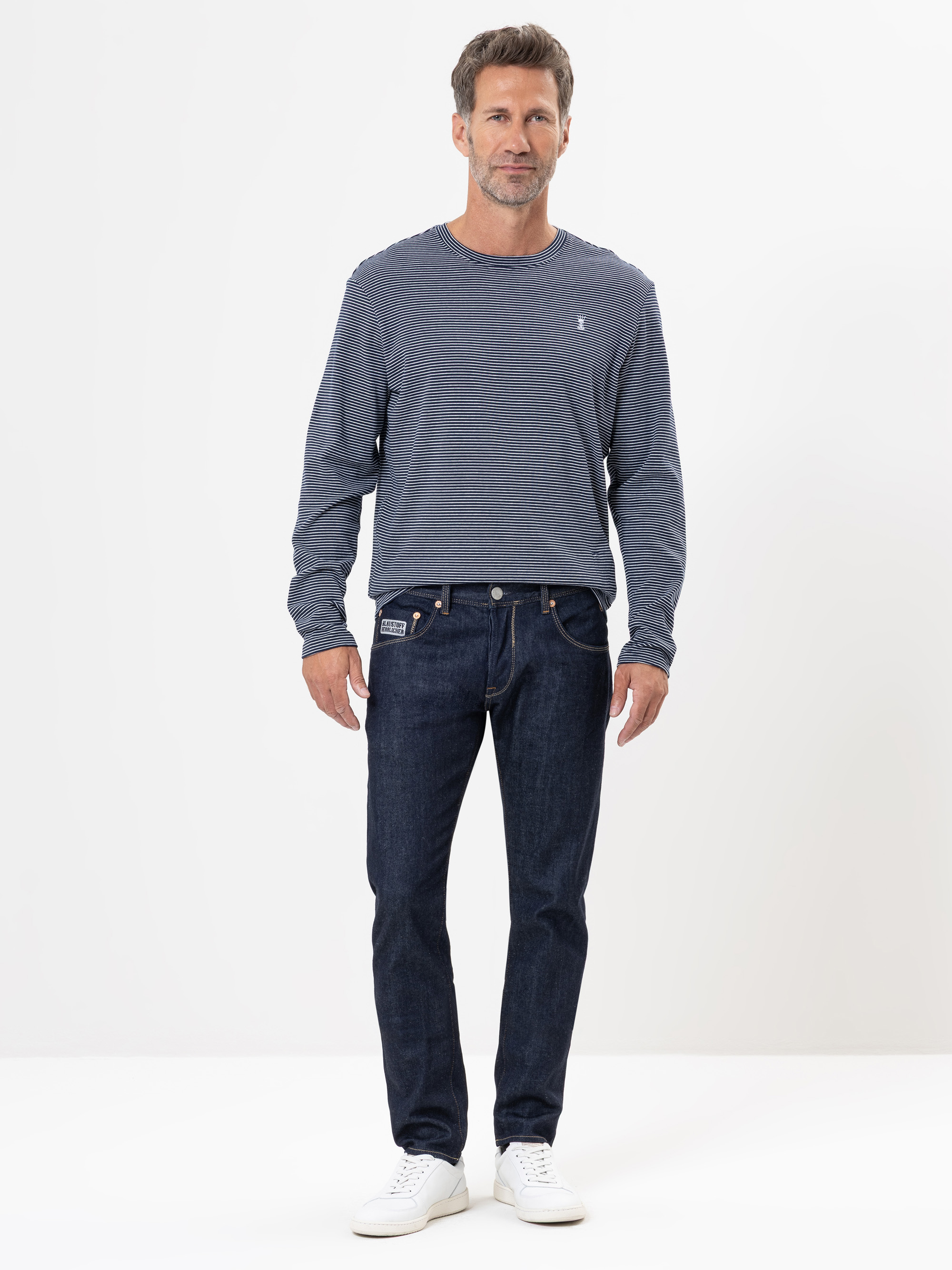 Ein Mann steht vor einem schlichten weißen Hintergrund und trägt ein langärmeliges, blau gestreiftes Hemd, dunkelblaue Jeans und weiße Turnschuhe. Er blickt nach vorne und hat einen neutralen Gesichtsausdruck.