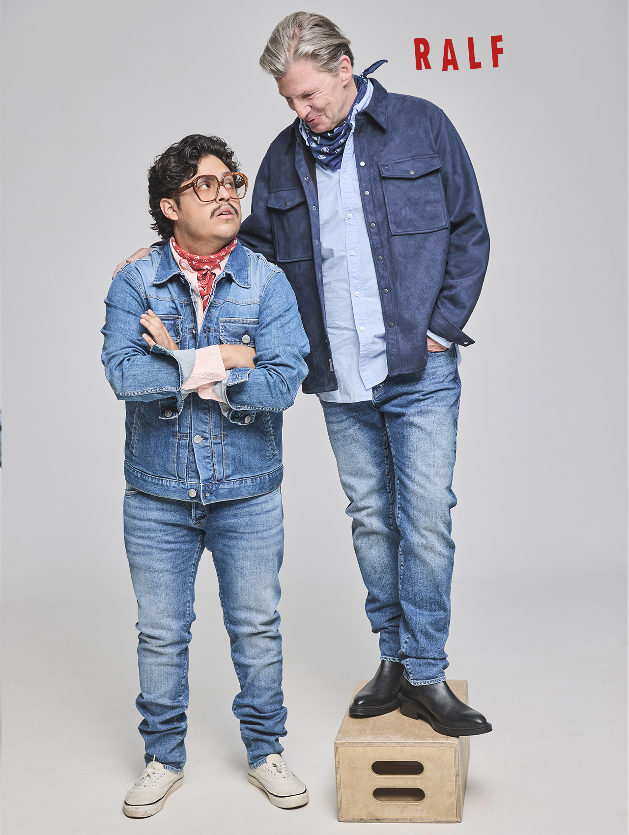 Zwei Personen posieren zusammen; die eine steht auf einer Kiste und schaut auf die andere herab, die mit verschränkten Armen dasteht. Beide tragen Jeanskleidung und Schals. Über ihnen steht in roter Schrift das Wort RALF auf einfarbigem Hintergrund.
