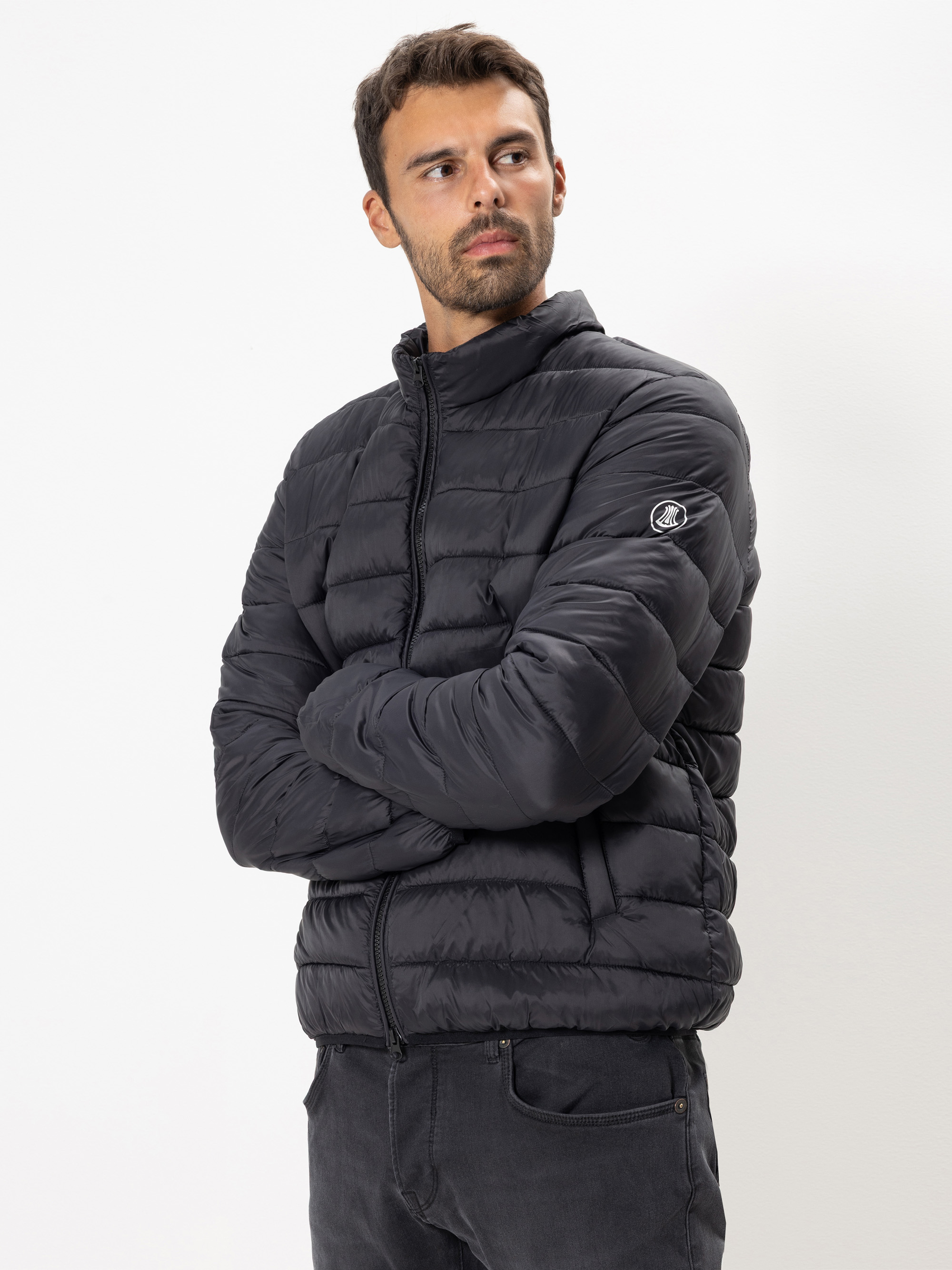 Ein Mann mit kurzen dunklen Haaren und einem Bart trägt eine schwarze Pufferjacke mit einem Reißverschluss und einem Logo auf dem Ärmel. Er steht mit verschränkten Armen vor einem schlichten weißen Hintergrund und blickt leicht nach links.