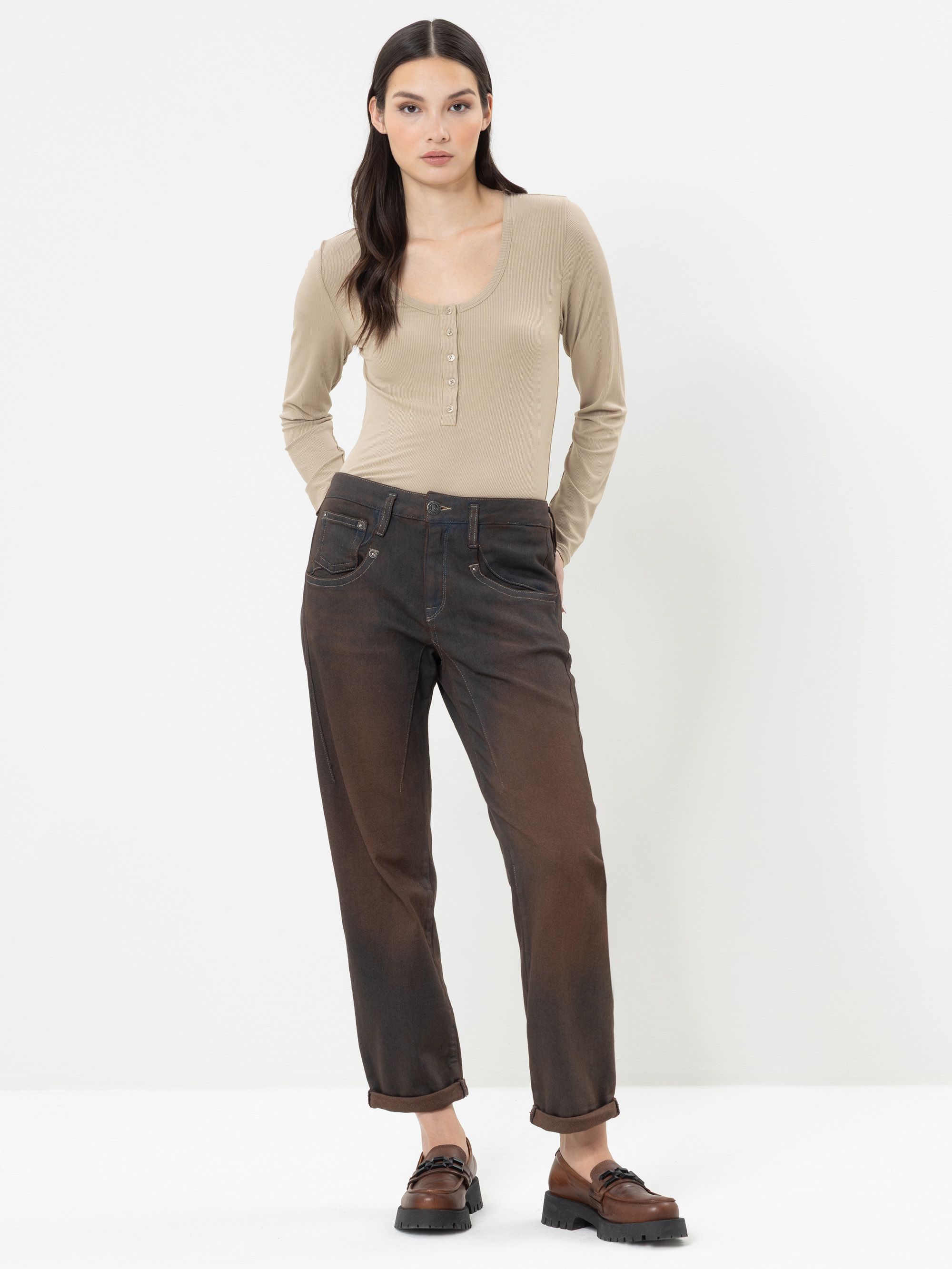 Vor einem weißen Hintergrund steht eine Frau, die ein beiges Langarm-Oberteil, eine ausgewaschene braune Jeans mit umgeschlagenen Bündchen und braune Slipper trägt.