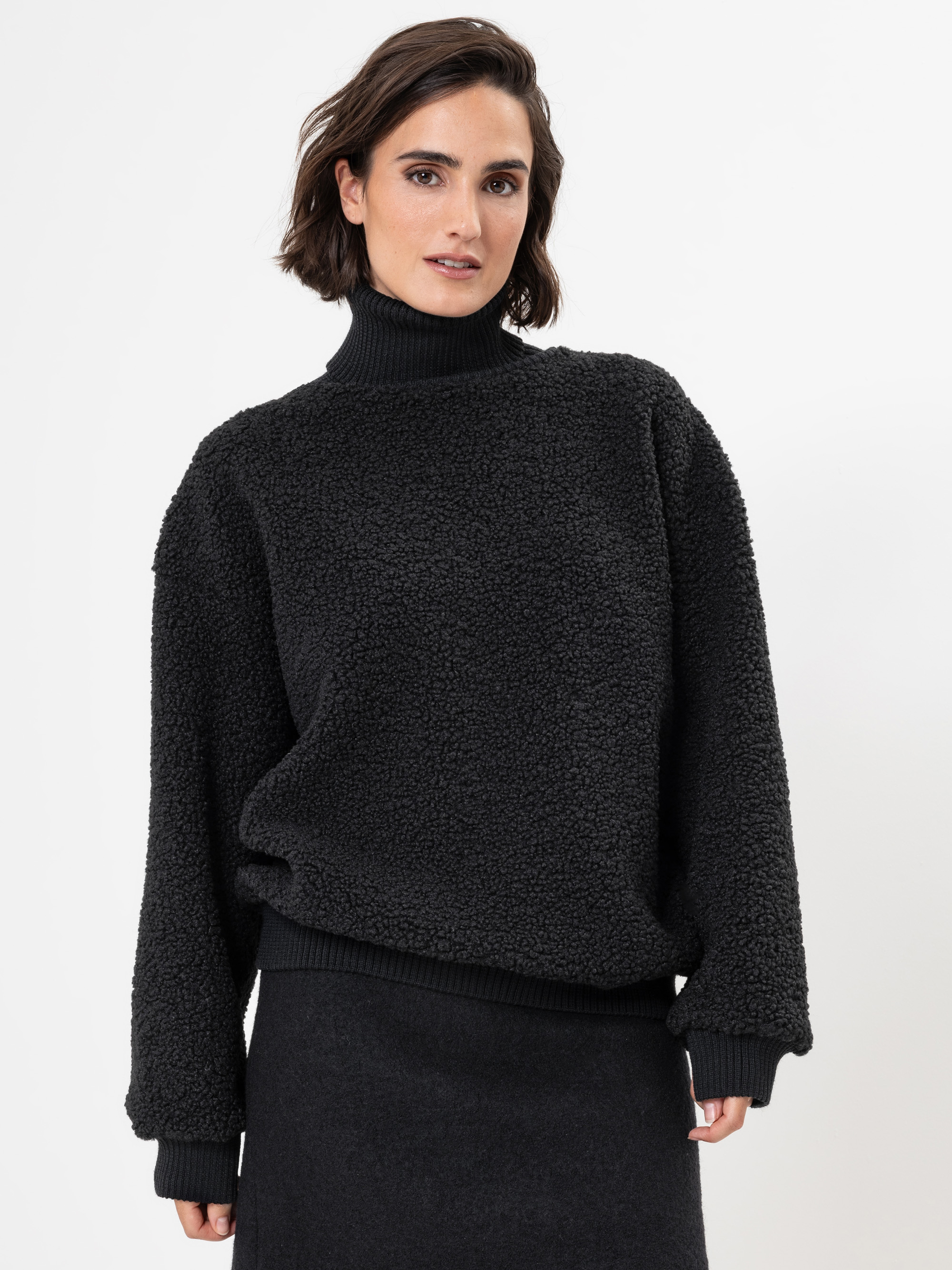 Eine Frau mit kurzen braunen Haaren trägt einen strukturierten schwarzen Rollkragenpullover und einen dazu passenden dunklen Rock und steht vor einem schlichten weißen Hintergrund.