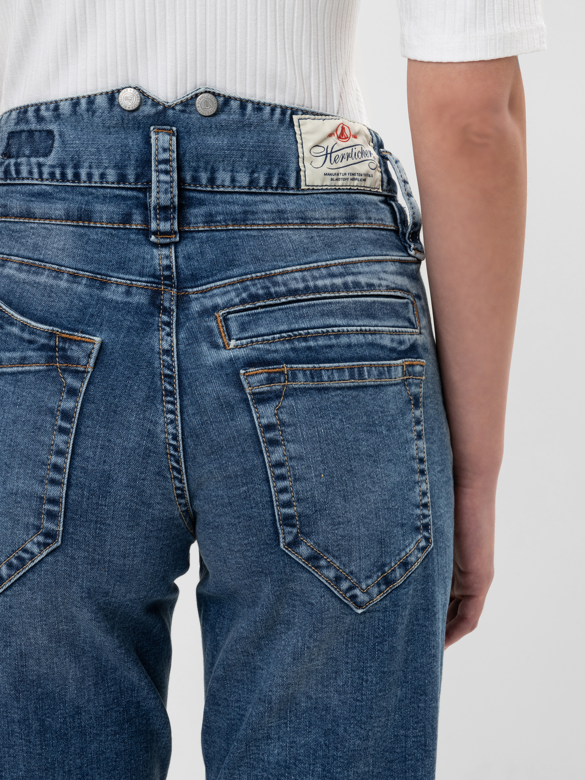 Eine Person trägt eine blaue High-Waisted-Jeans mit doppelten Gesäßtaschen und ein weißes geripptes Oberteil. Die Jeans hat ein sichtbares Markenetikett am Bund.