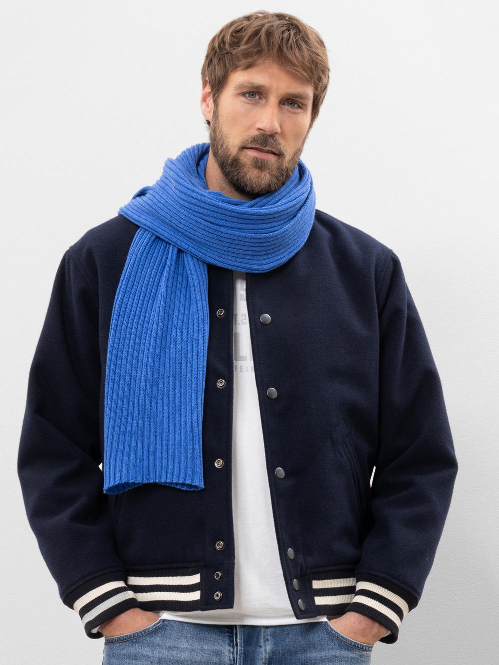 Ein Mann mit hellbraunem Haar und Bart trägt eine dunkle Sportjacke, ein weißes Hemd, blaue Jeans und einen hellblauen Strickschal. Er steht vor einem schlichten weißen Hintergrund und hat die Hände in den Jackentaschen.