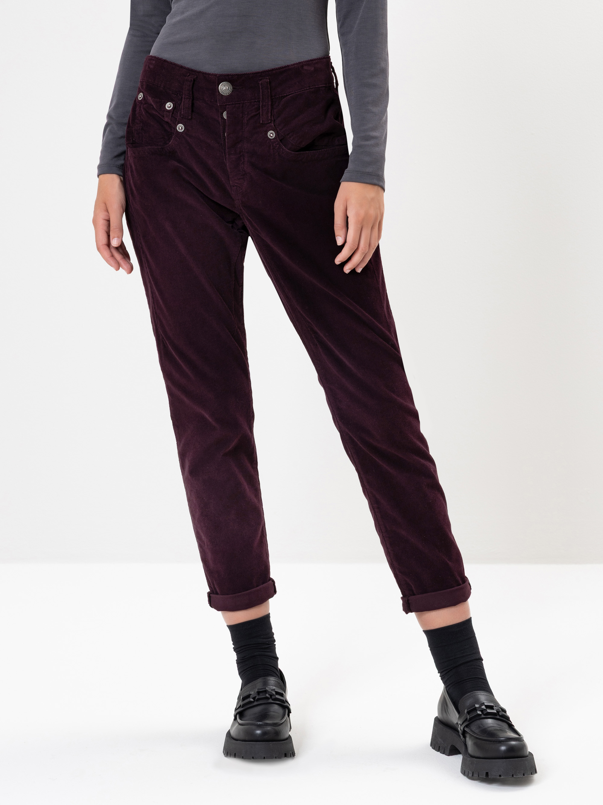 Eine Person, die eine dunkelrote Cordhose, ein graues, langärmeliges Oberteil, schwarze Socken und schwarze Halbschuhe trägt, steht mit einem leicht angewinkelten Bein vor einem schlichten weißen Hintergrund.