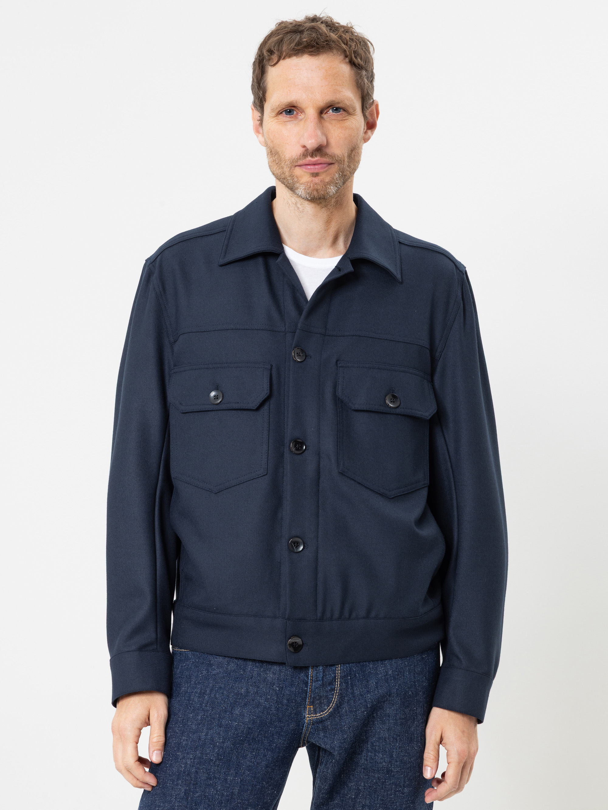 Ein Mann, der eine marineblaue Jacke mit Knöpfen und Pattentaschen über einem weißen Hemd und blauen Jeans trägt, steht mit dem Blick nach vorne vor einem schlichten weißen Hintergrund.