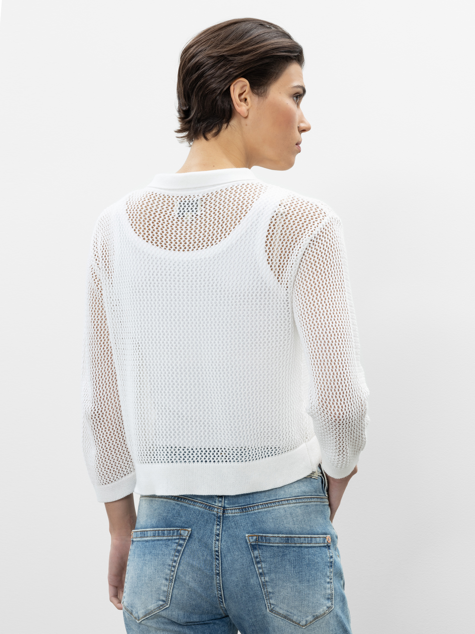 Eine Person mit kurzen braunen Haaren trägt einen weißen, langärmeligen Netzpullover über einem weißen Tanktop und eine blaue Jeans. Sie blickt von der Kamera weg auf einen schlichten, hellen Hintergrund.