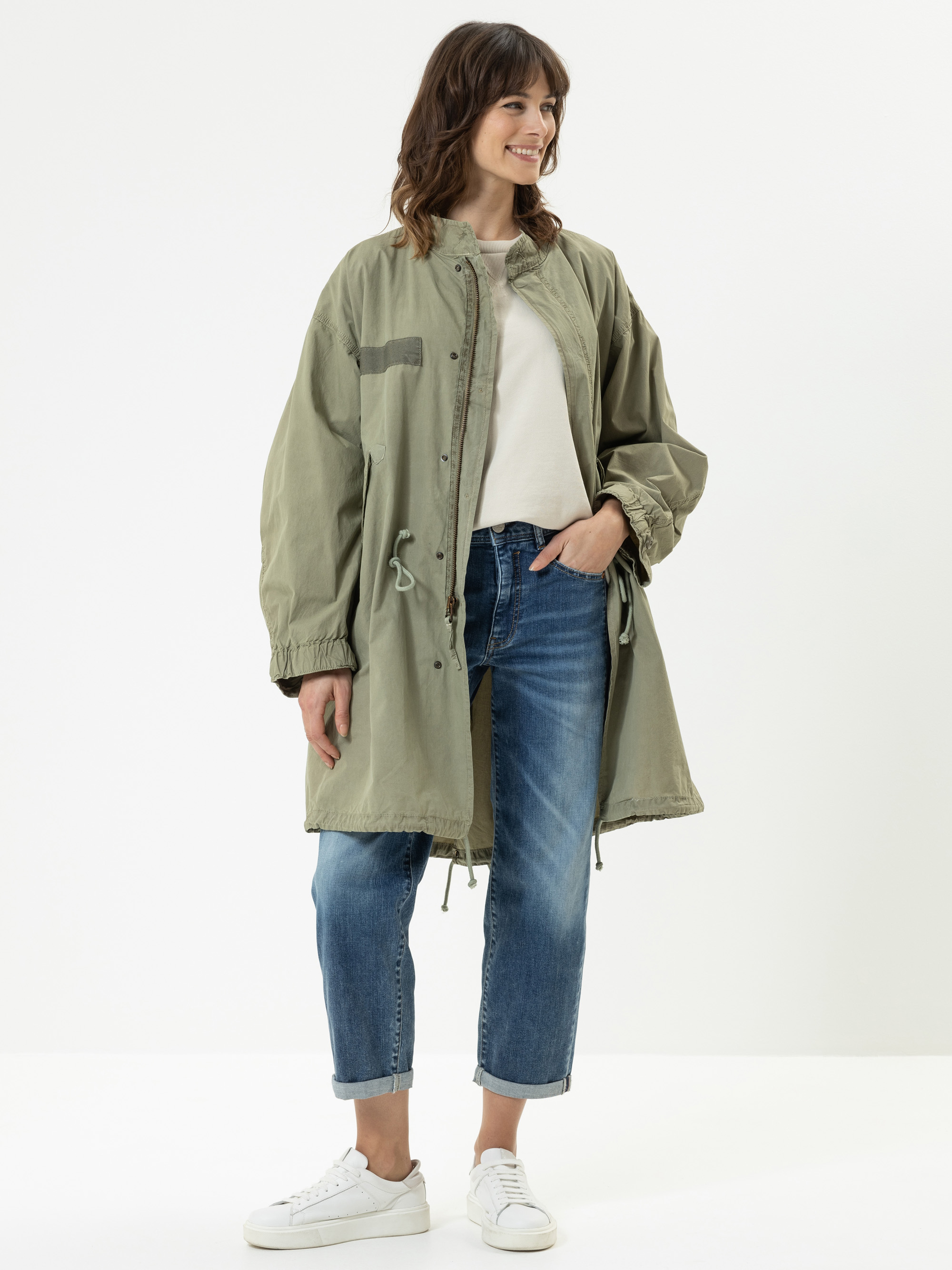 Eine Frau steht lächelnd da und trägt eine hellgrüne, übergroße Jacke über einem weißen Oberteil, blaue Jeans mit Bündchen und weiße Turnschuhe. Sie hat gewelltes braunes Haar und ihre linke Hand ruht in ihrer Jeanstasche. Der Hintergrund ist ein