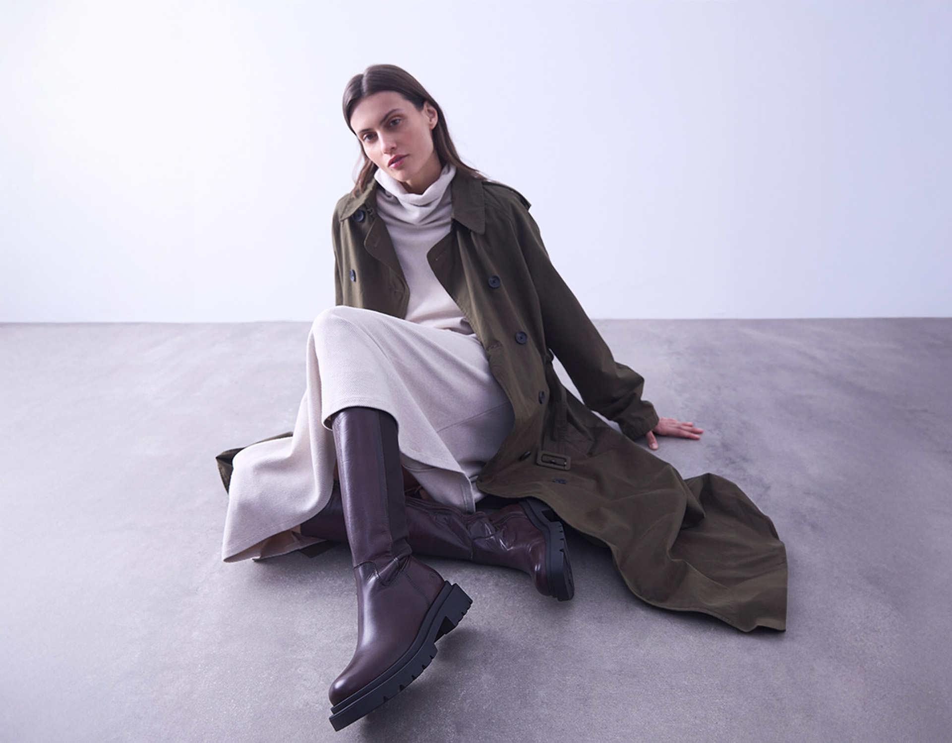Eine Frau in einem langen olivgrünen Trenchcoat, einem cremefarbenen Rollkragenkleid und dunkelbraunen, kniehohen Stiefeln sitzt auf einem Betonboden und lehnt sich mit einer Hand auf dem Boden an eine schlichte, helle Wand.