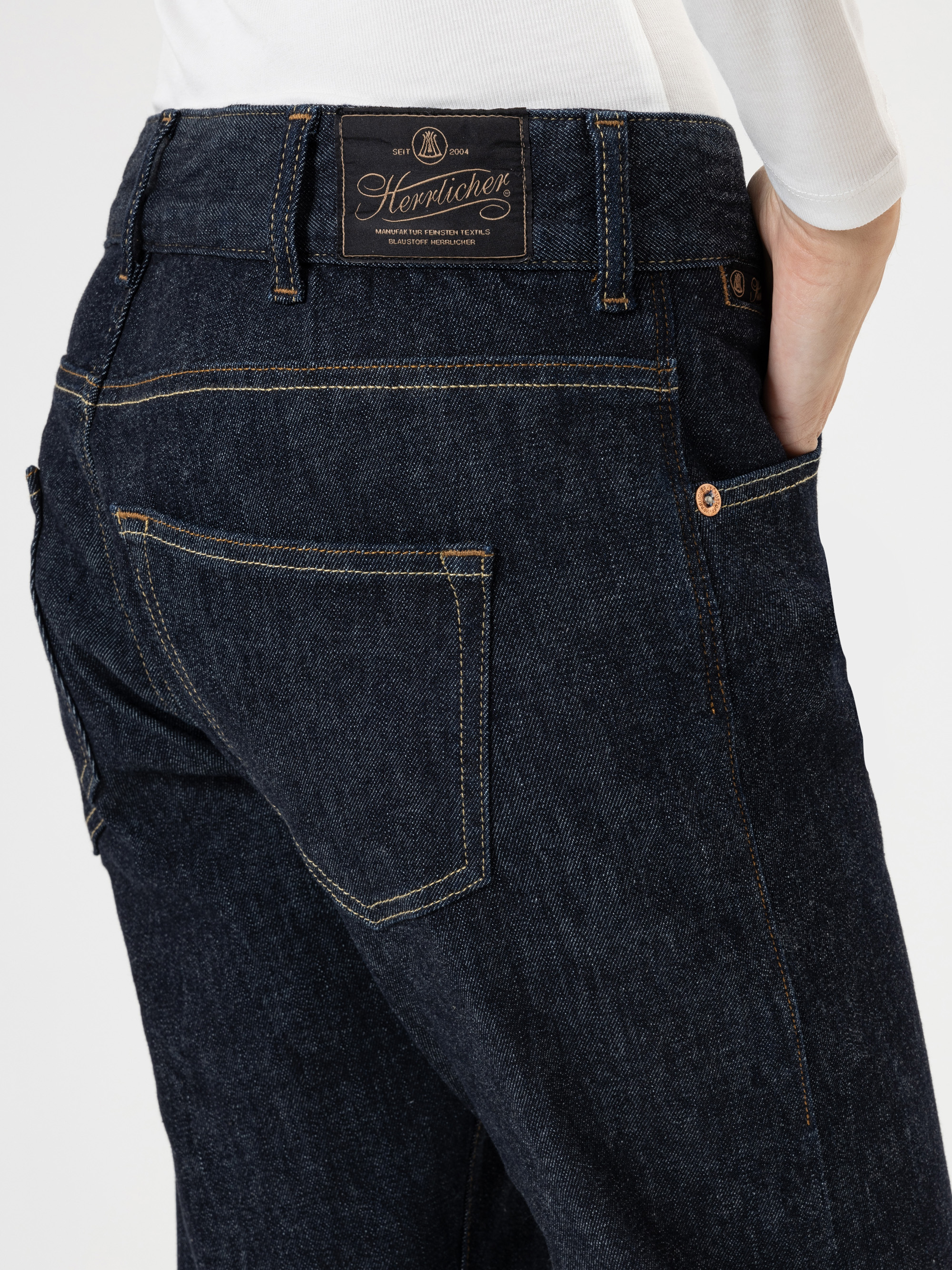 Eine Person trägt eine dunkelblaue Jeans mit Kontrastnähten, einer sichtbaren Gesäßtasche und einem Herrlicher-Markenetikett am Bund. Die Hand der Person befindet sich in der Gesäßtasche, und sie trägt ein weißes Oberteil.