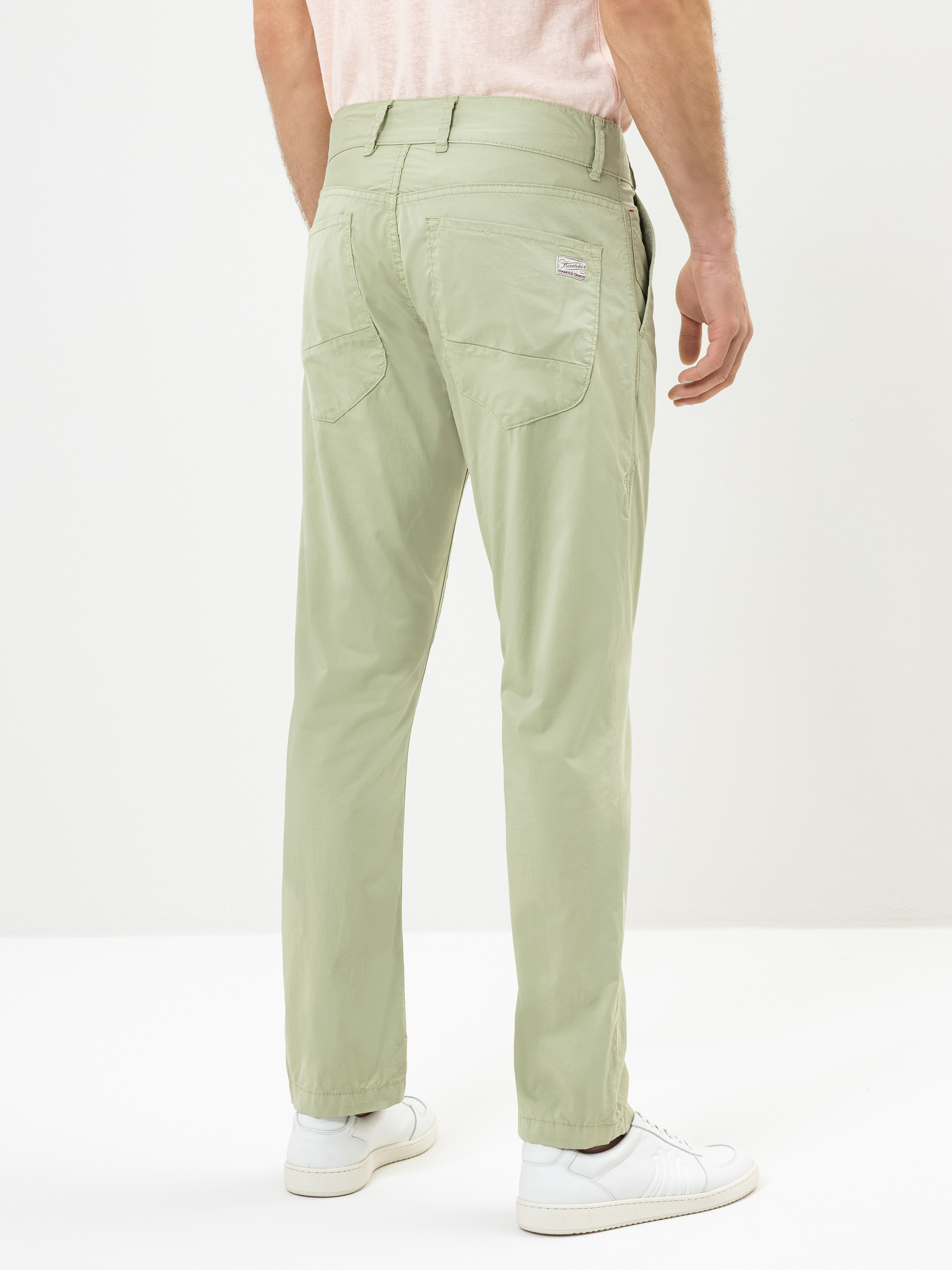 Ein Mann steht mit dem Gesicht zur Wand, er trägt eine hellgrüne Hose, ein hellrosa Hemd, das in die Hose gesteckt ist, und weiße Turnschuhe. Der Hintergrund ist schlicht und weiß.